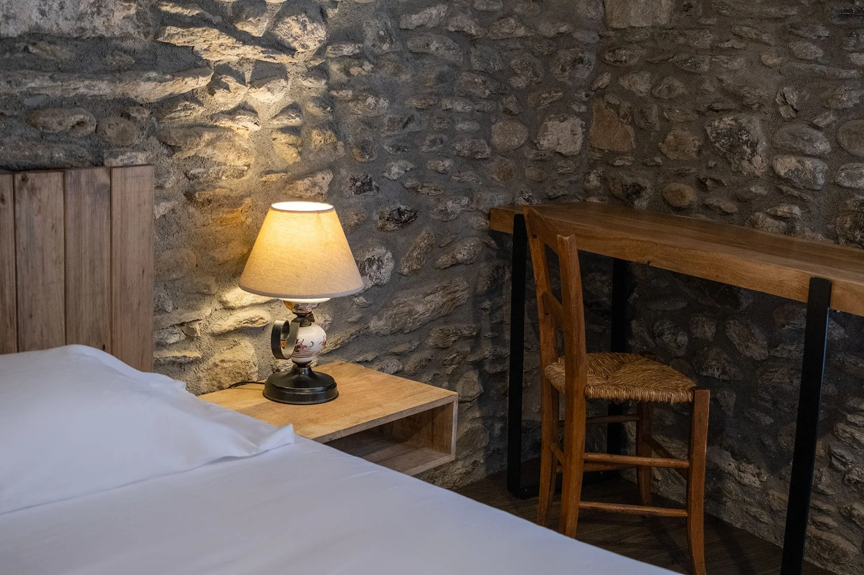 Habitación con pared de piedra, cama con sábanas blancas, lámpara encendida sobre mesa de noche de madera, escritorio de madera y silla de madera con respaldo de mimbre.