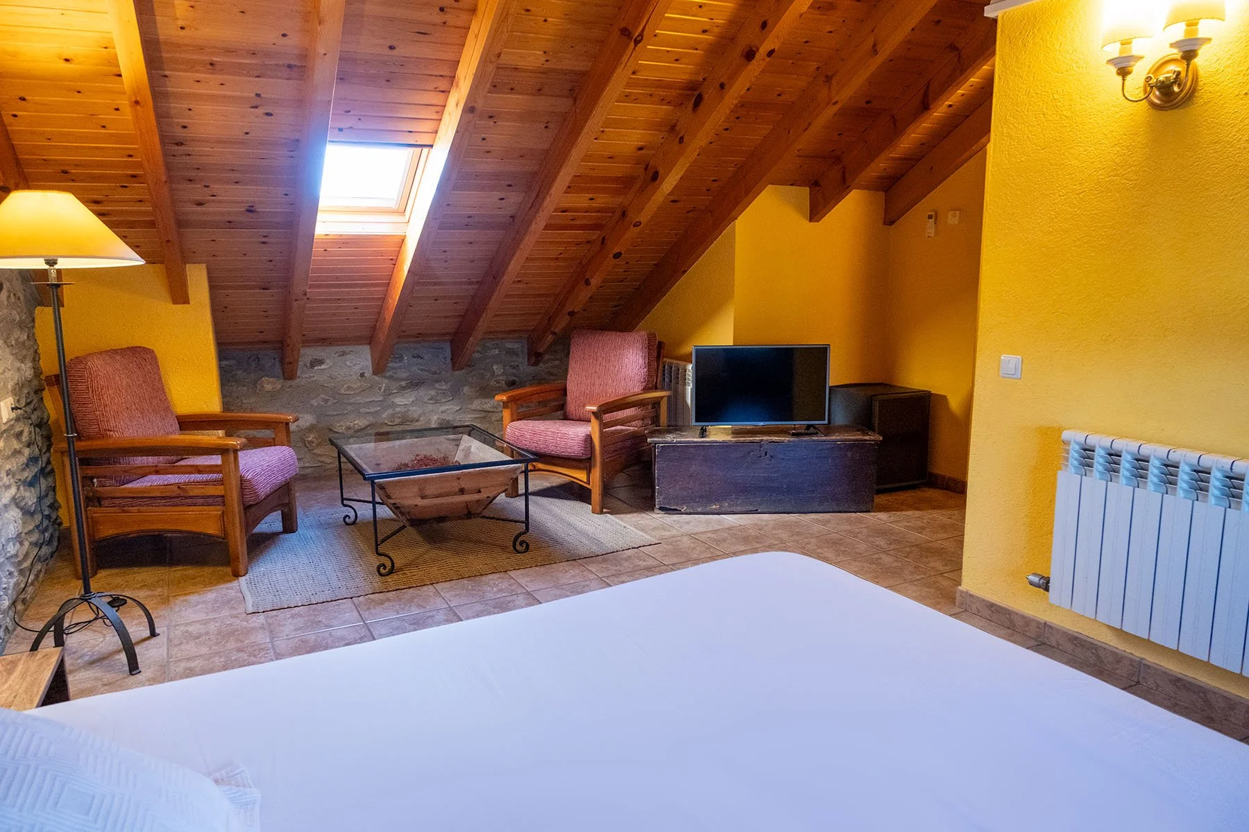 Interior de habitación con techo de madera, ventanas en el techo, sillones de madera con cojines, televisión en una mesa de madera, paredes amarillas y suelo de baldosas.