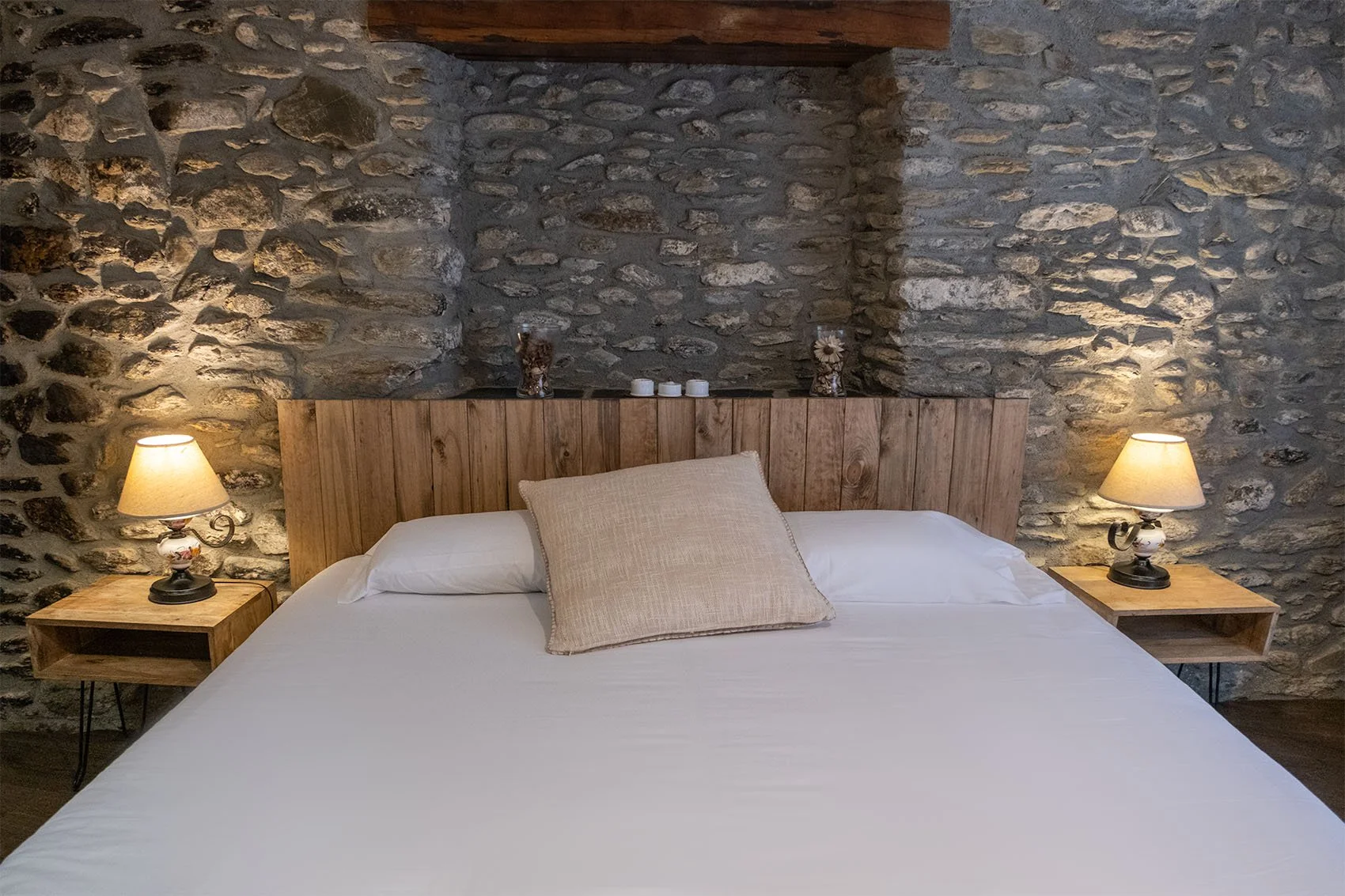 Cama con cabecera de madera en una habitación con pared de piedra, con dos lámparas de noche encendidas y un cojín en la cama.