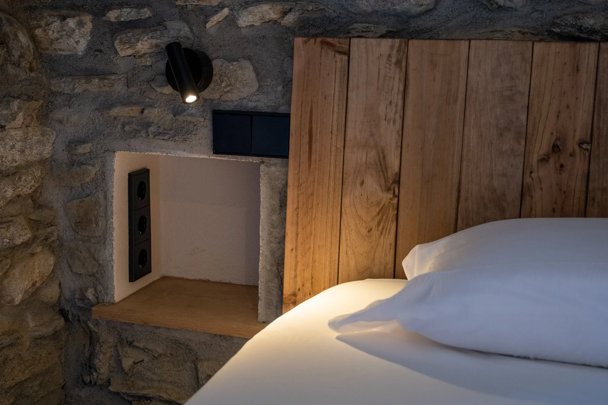Imagen de una habitación con pared de piedra, un cabecero de madera sobre la cama con sábanas blancas, y una lámpara negra en la pared