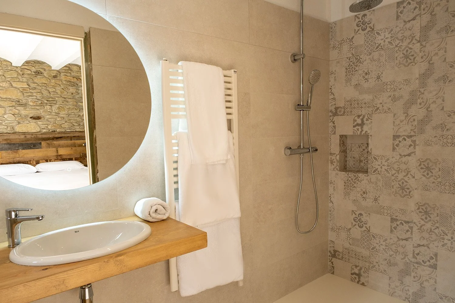 Baño moderno con lavabo, espejo redondo, toallas blancas en radiador, ducha con ducha de mano y pared decorativa con azulejos de patrones geométricos y florales.