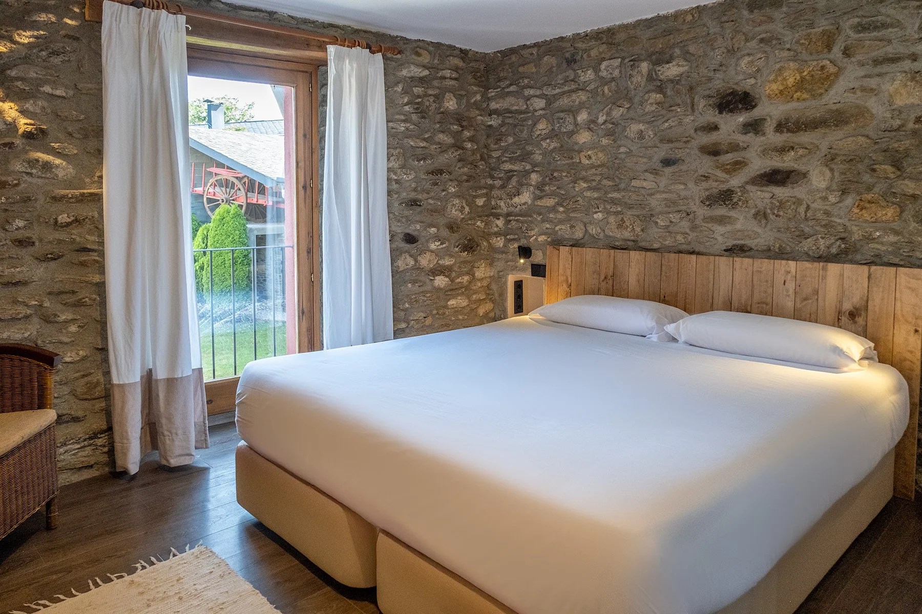 Habitación con paredes de piedra, cama grande con cabecera de madera, cortinas blancas, y puerta de vidrio que da a un balcón con vista de un jardín y estructura de madera exterior.