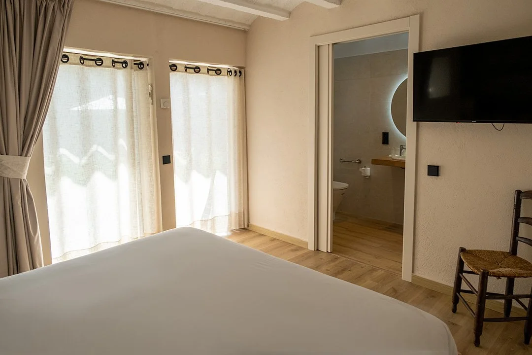 Habitación con cama, cortinas blancas, ventanas, puerta al baño, televisor y silla de madera.