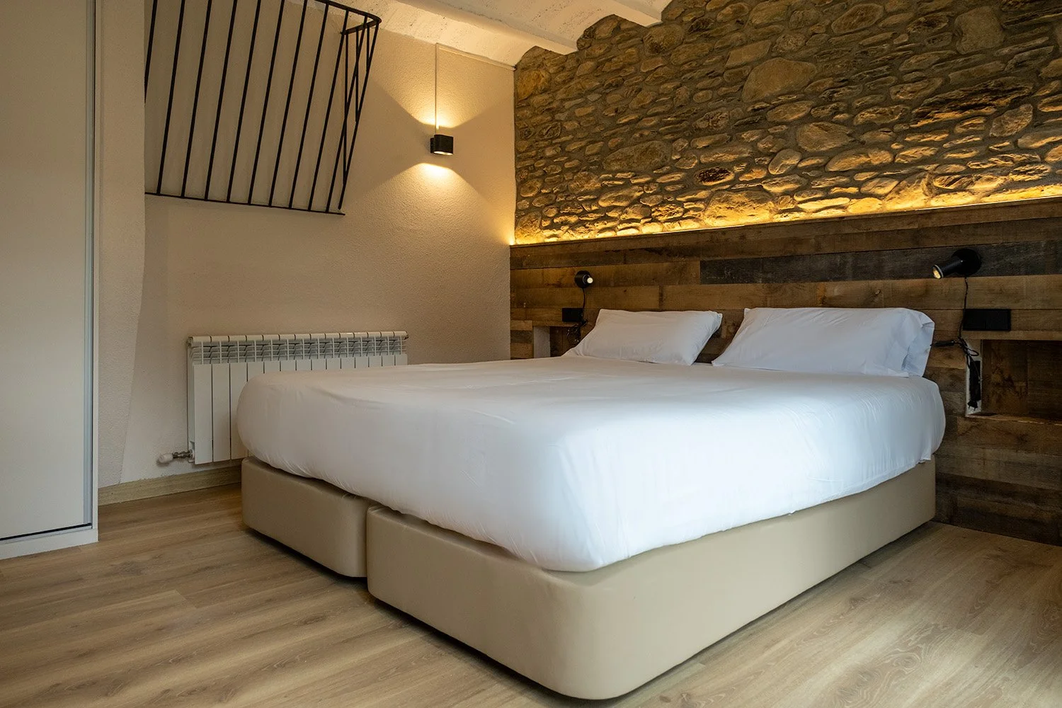 Habitación moderna con cama grande, paredes de piedra y madera, iluminación suave y detalles decorativos en tonos neutros.