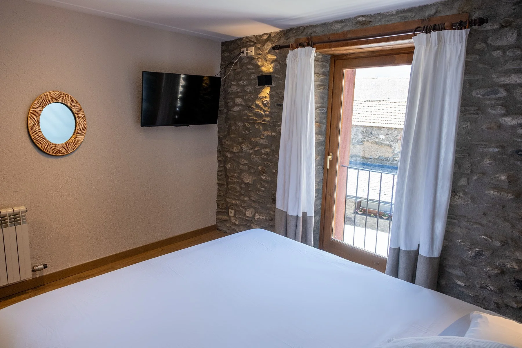 Habitación con pared de piedra, ventana con cortinas blancas y vistas a tejados de otras casas.