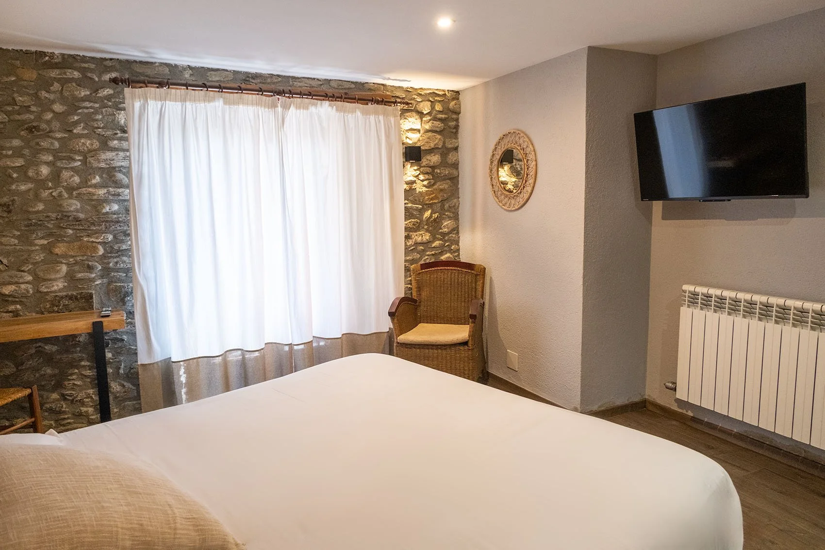 Habitación con pared de piedra, cortinas blancas, silla de mimbre, televisor en la pared y un radiador.