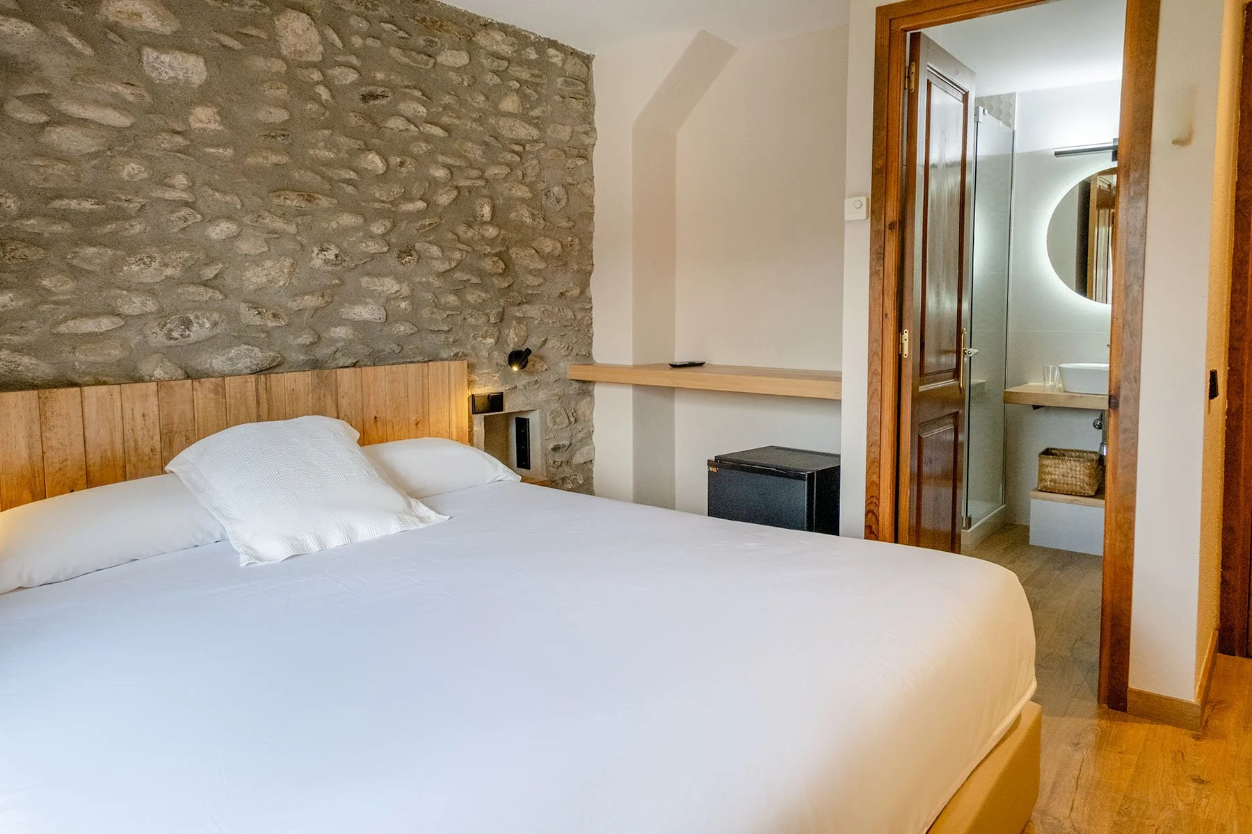 Habitación con cama grande, cabecera de madera, pared de piedra y acceso a baño con lavabo y espejo redondo