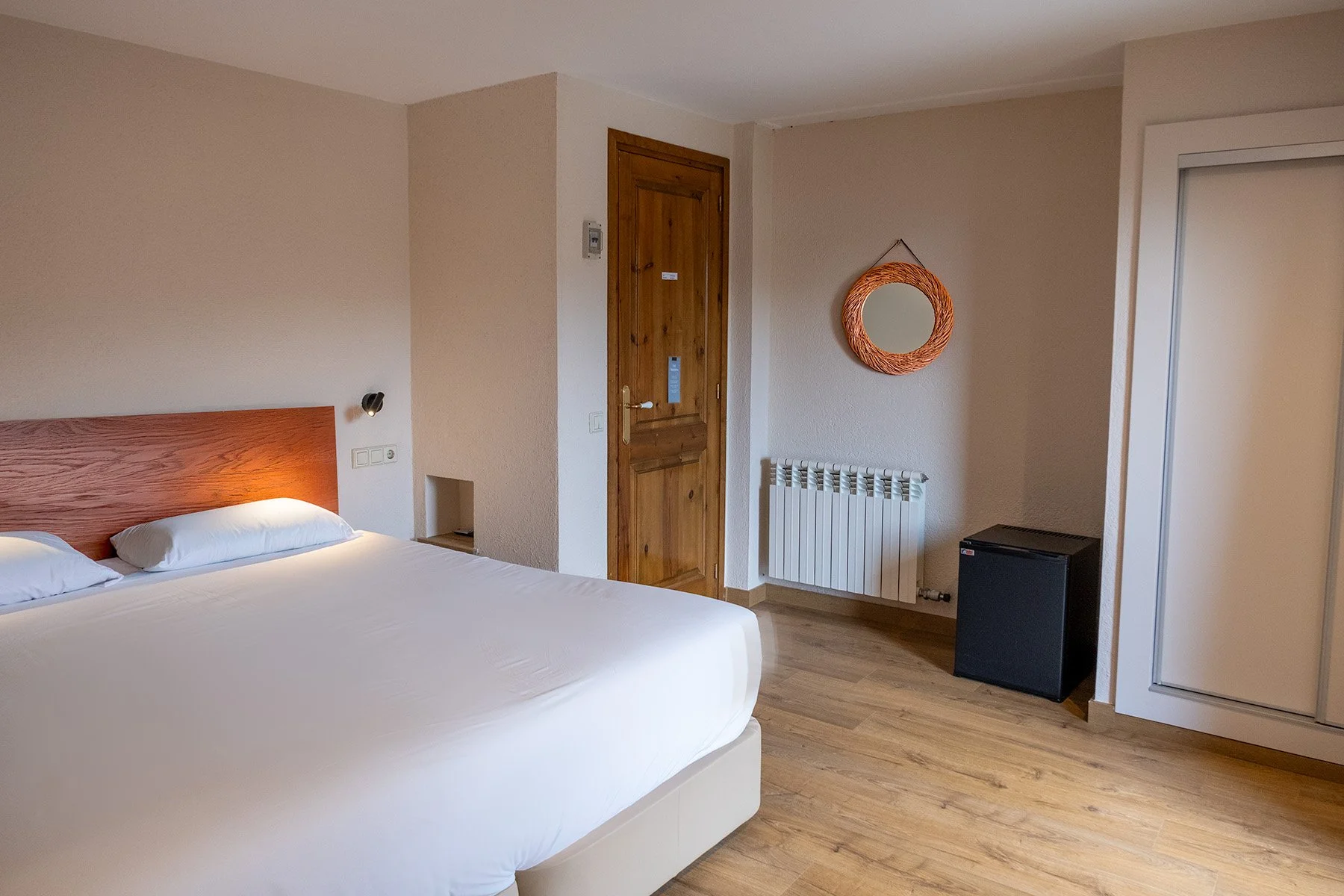 Habitación de hotel con cama doble, pared beige, espejo redondo de mimbre, radiador, mini refrigerador y armario con puertas blancas