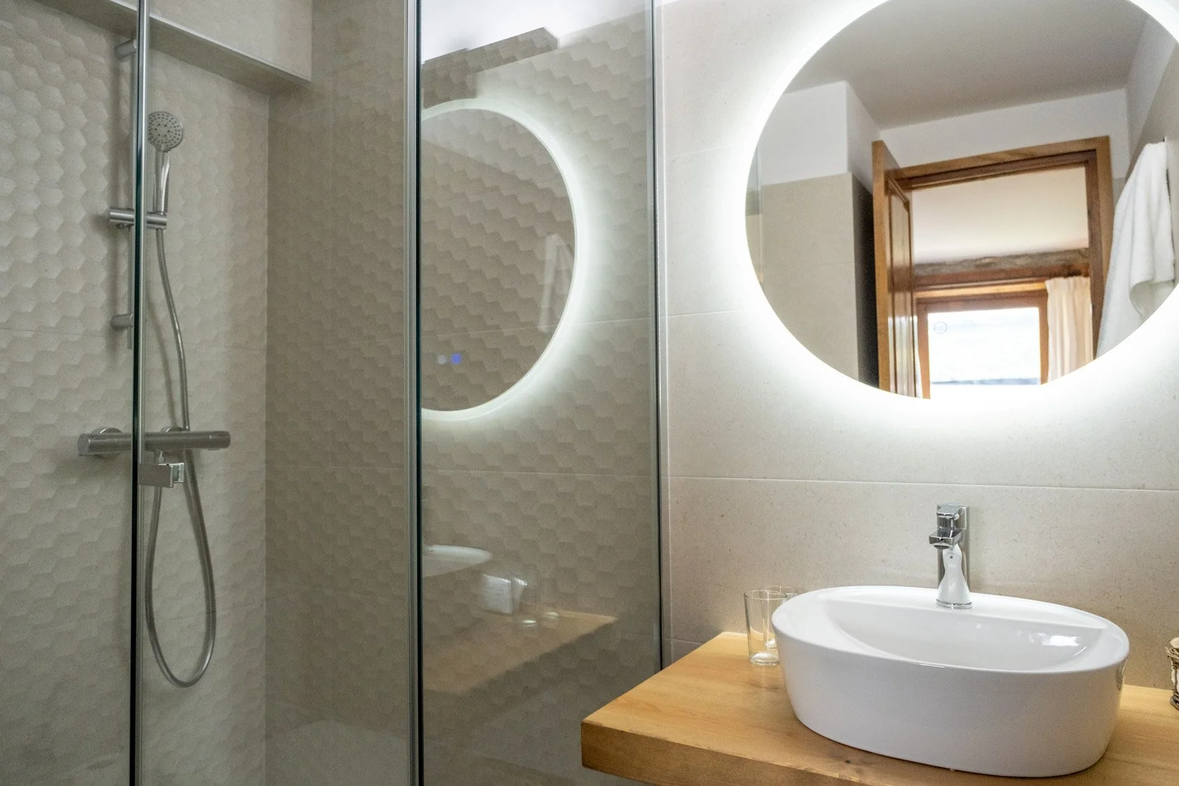 Lavabo moderno con espejo con luz de fondo, ducha con puerta de cristal y pared de azulejos texturizados en un baño.