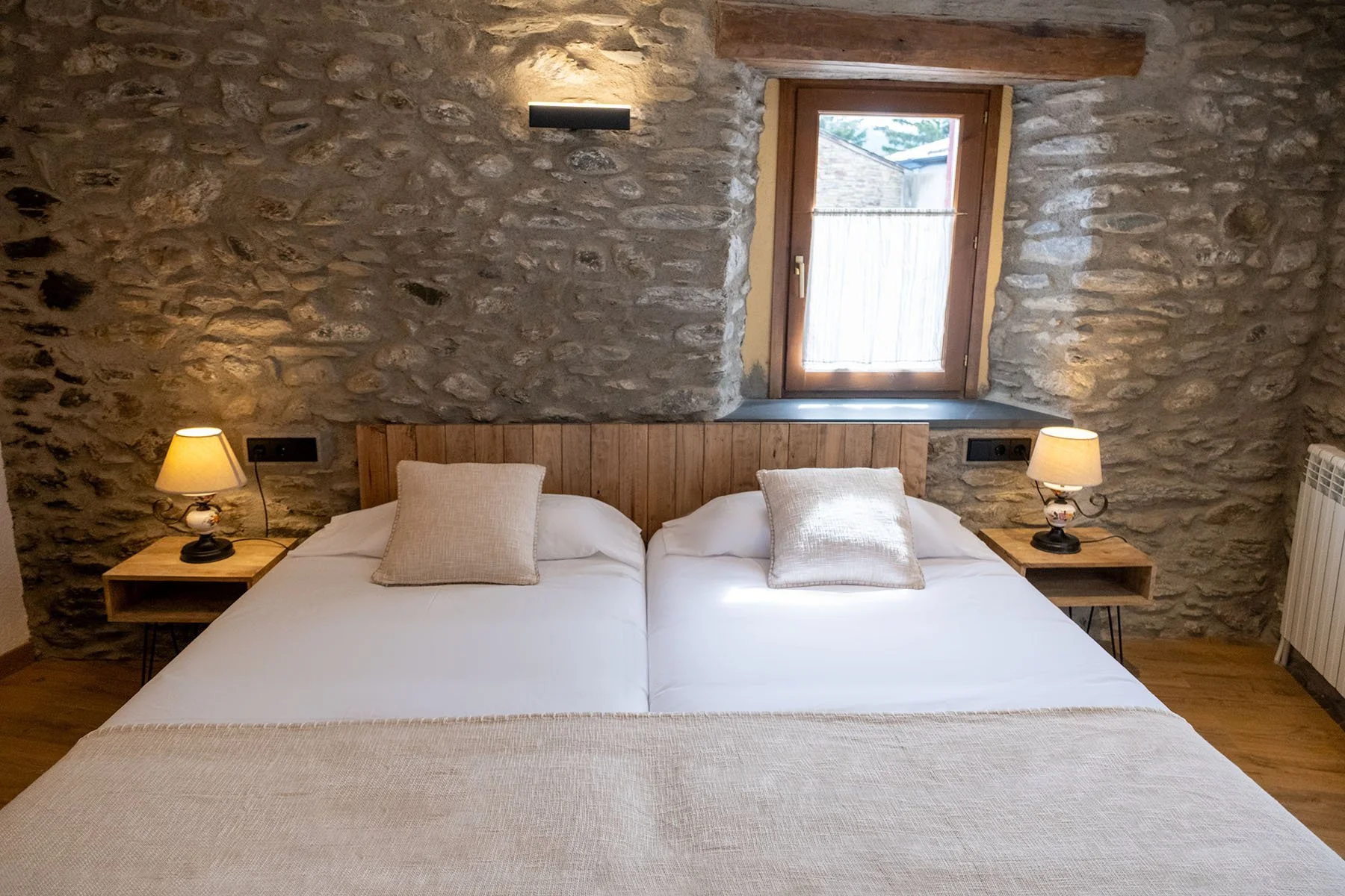 Habitación con paredes de piedra, dos camas juntas con blancas sábanas, almohadas beige, cabecera de madera, dos mesas de noche con lámparas, una ventana con cortina de tela ligera, piso de madera.