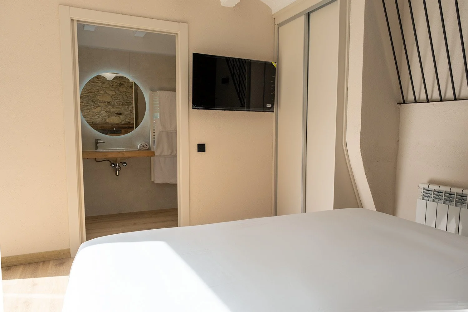 Habitación con cama, televisión en la pared, baño visible con espejo y lavabo, y una escalera con barandal negro.