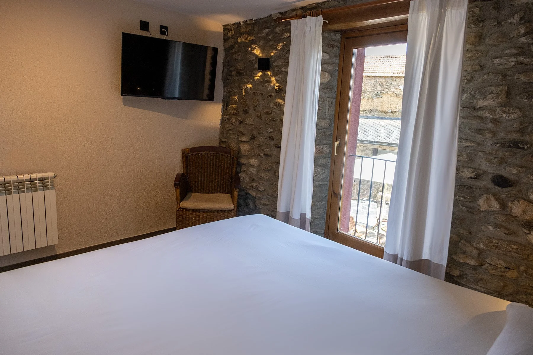 Habitación con pared de piedra, ventana con cortinas blancas, televisor de pantalla plana, silla de mimbre, cama con sábanas blancas, radiador y vista exterior de casas antiguas.