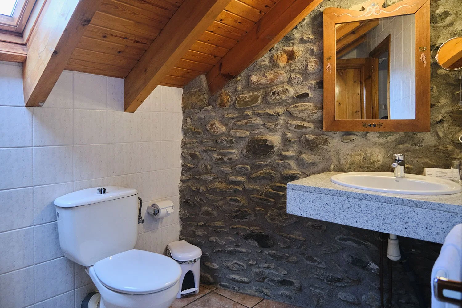 Baño con pared de piedra, lavabo de encimera de granito, espejo de madera y sanitario blanco, en un espacio con techo de madera y muro de azulejo.