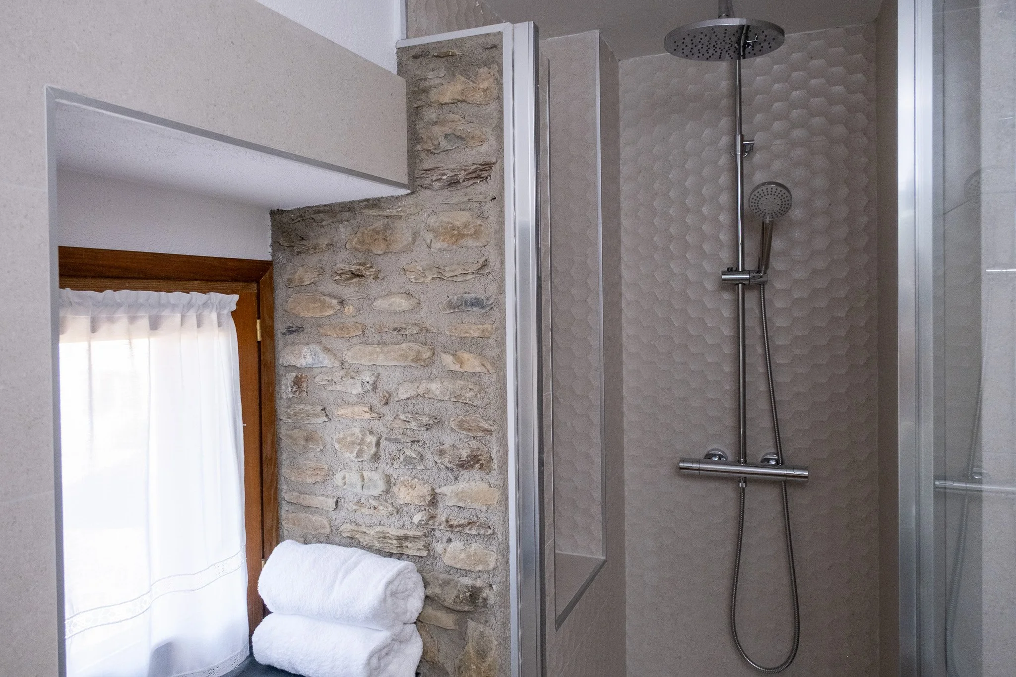 Ducha moderna en un baño con paredes de azulejo, una pared de piedra en un espacio abierto, y toallas blancas apiladas debajo de una ventana con cortina blanca.