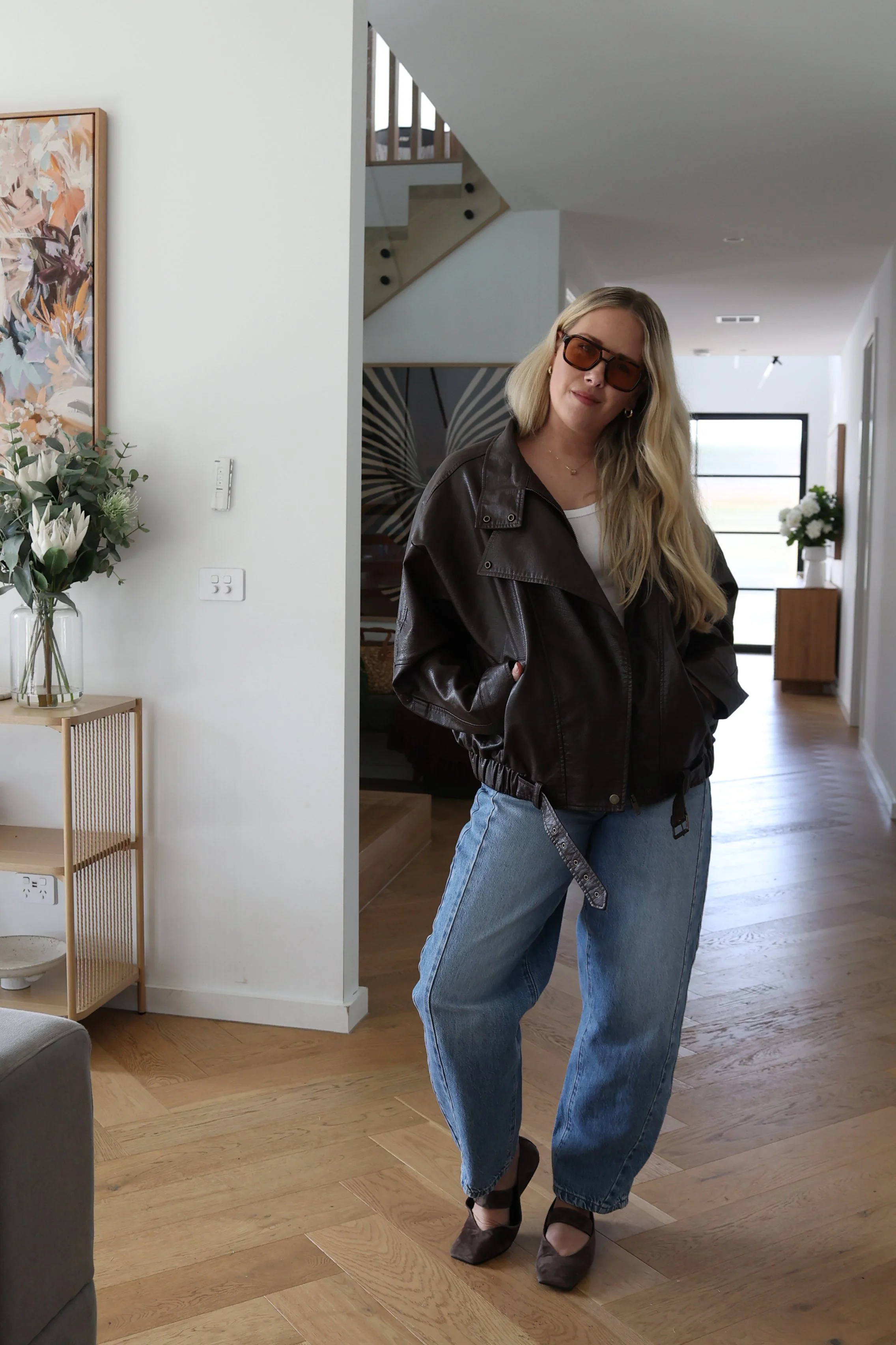 A woman with long blonde hair, wearing sunglasses, a brown leather jacket, white shirt, blue jeans, and high-heeled shoes, standing in a modern, well-lit living room with wooden floors, a small table with flowers, and artwork on the wall.