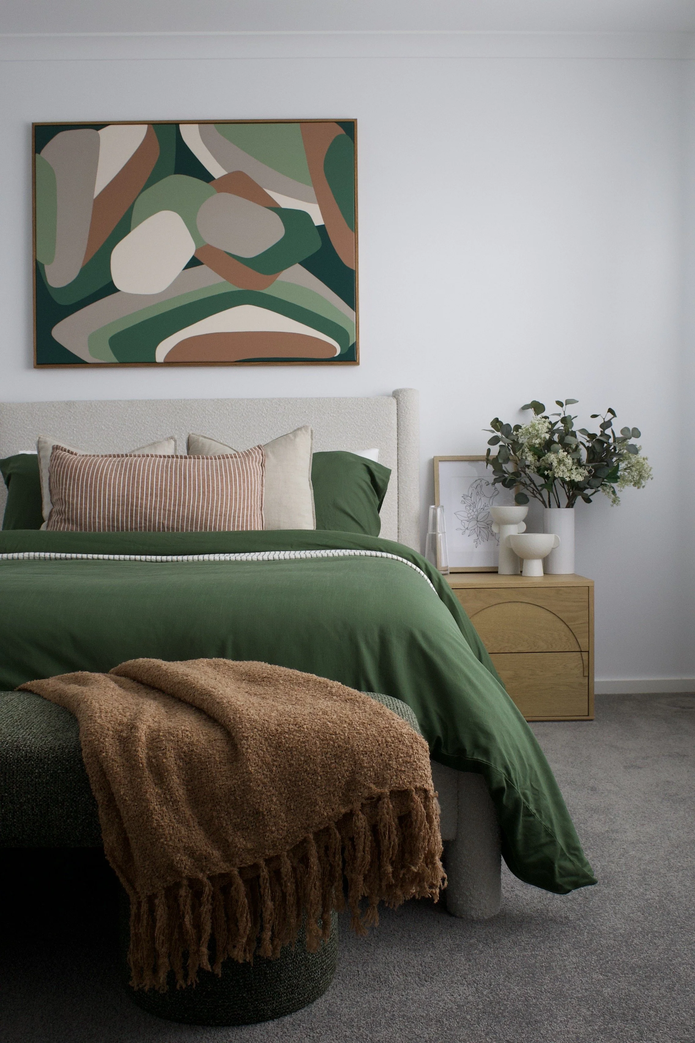 A bedroom scene featuring a bed with green bedding, beige pillows, and a striped throw blanket. A tan fringed blanket is draped over a woven ottoman at the foot of the bed. To the right of the bed is a wooden nightstand with a white vase holding gree