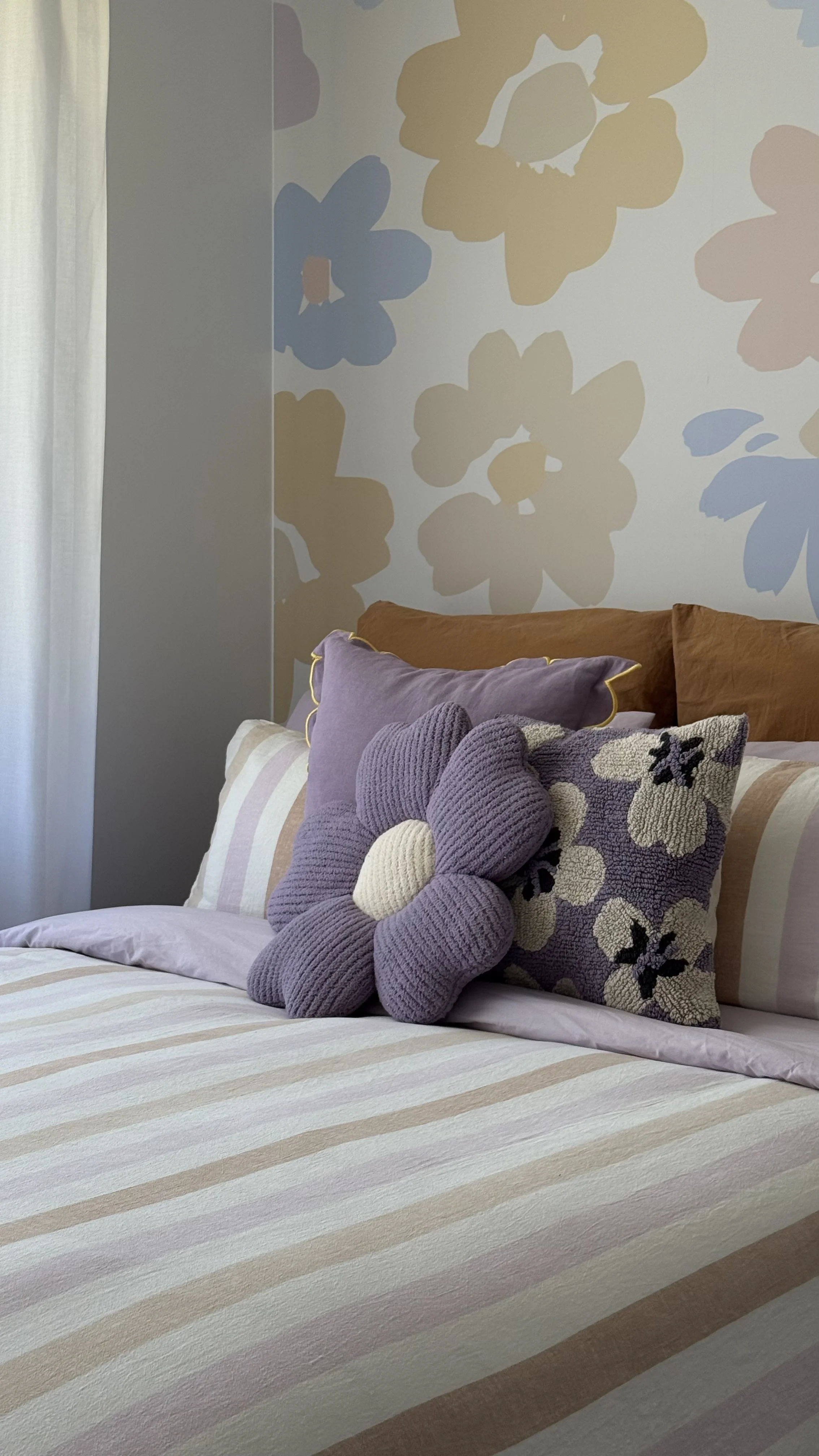 A bedroom bed with striped bedding, purple and floral pillows, three plush stuffed animals, and a large floral wallpaper on the wall behind.