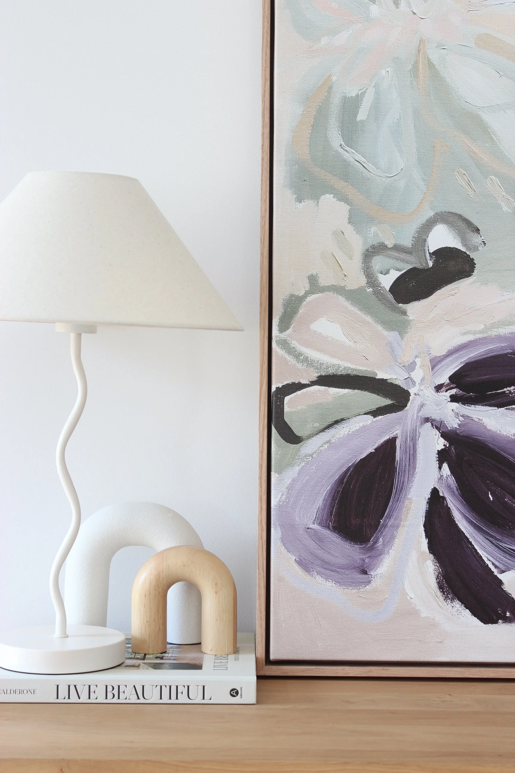A minimalist interior decor scene featuring a white table lamp with a curved stem, abstract wooden sculptures, and a large framed floral painting on a wooden surface.