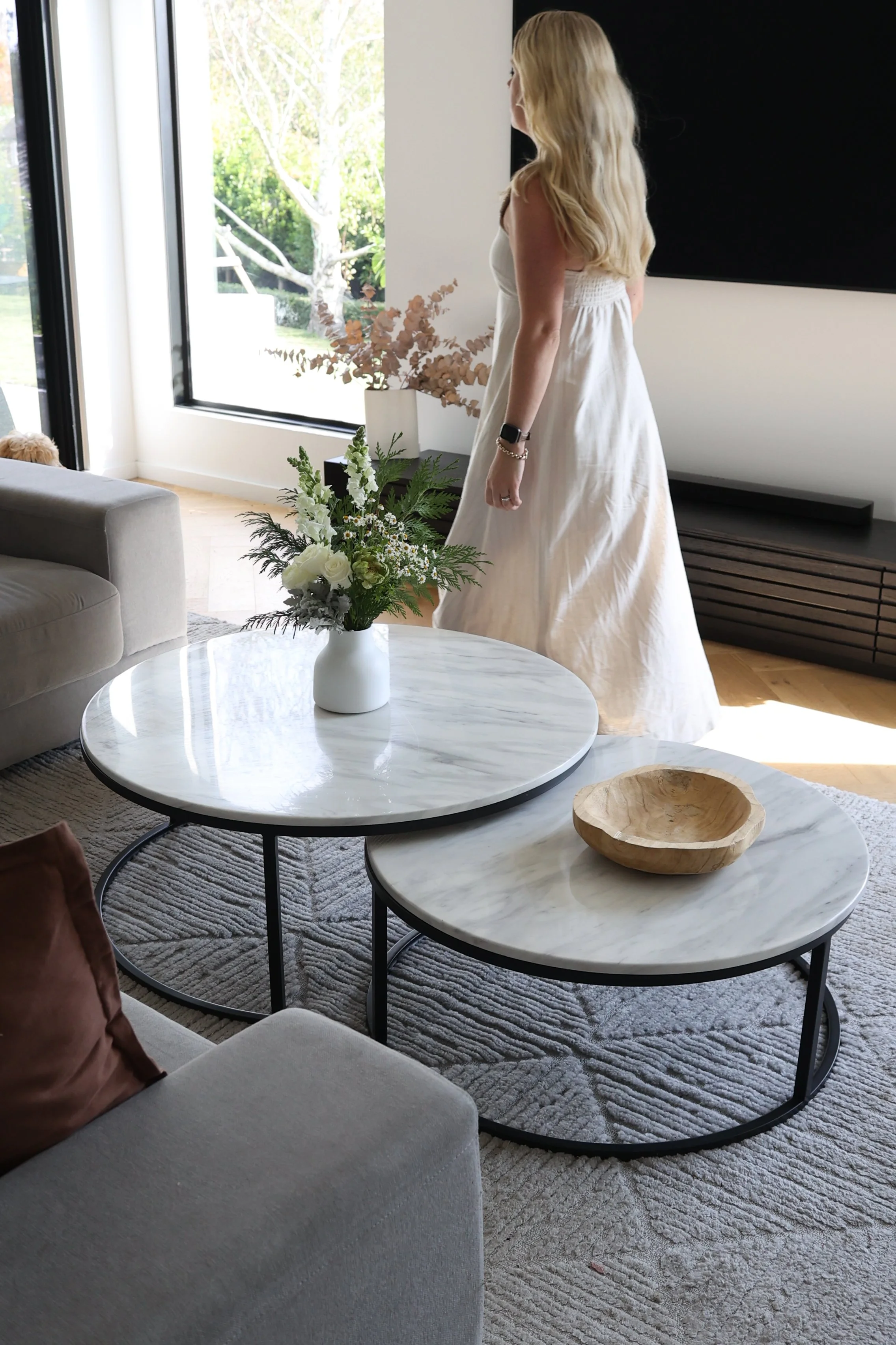 A woman with long blonde hair in a white dress standing in a modern living room next to a window with a view of trees outside, with a flower arrangement on the table in front of her.