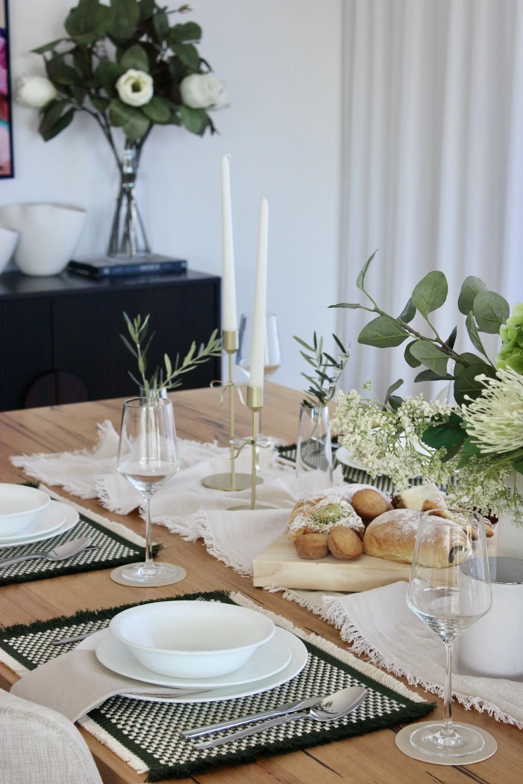 A dining table set for a meal with white plates, silverware, napkins, wine glasses, and a centerpiece of greenery and flowers. There are candles and a breakfast tray with bread and pastries. In the background, decorative vases and books are on a blac