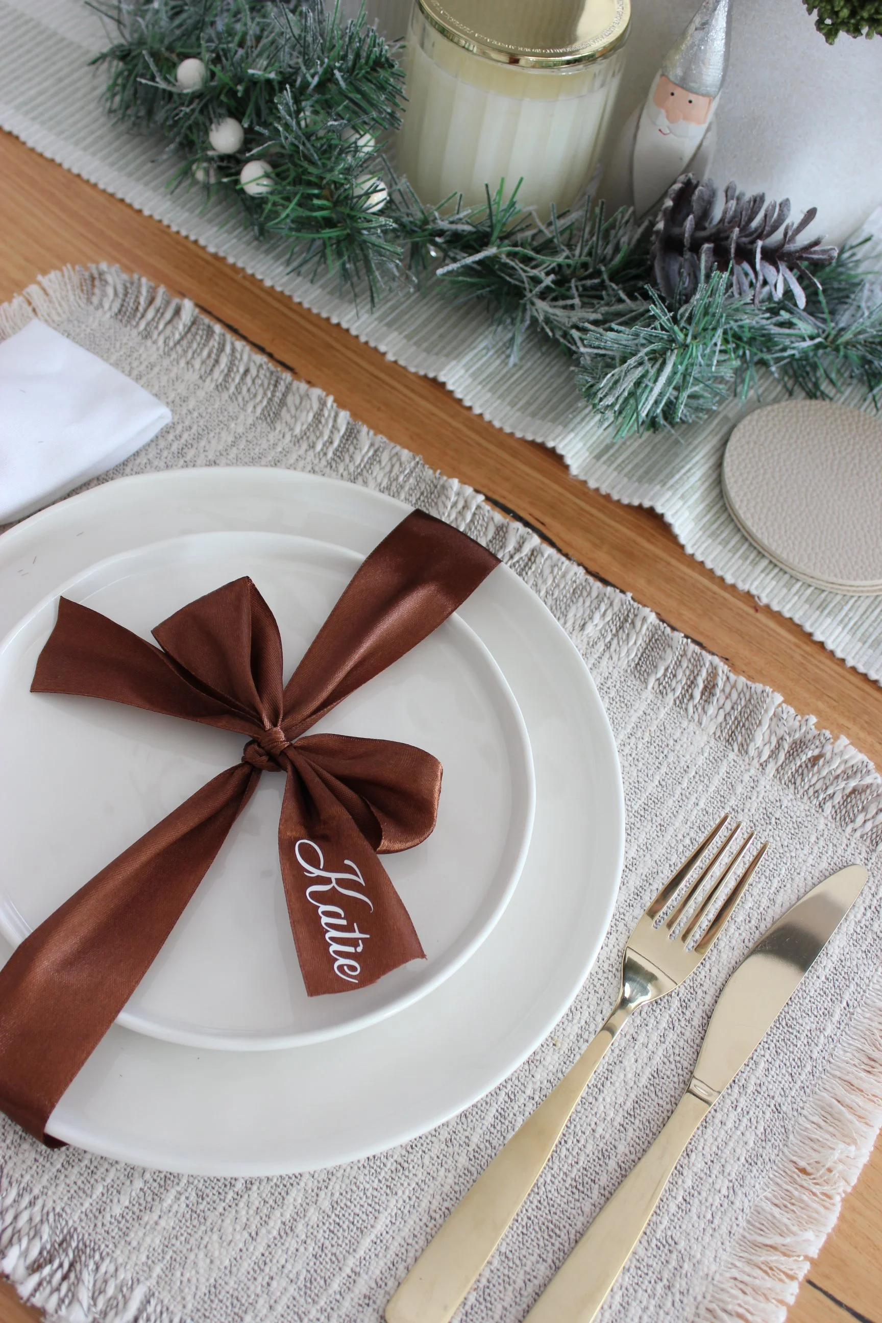 A table setting with a white plate decorated with a brown ribbon tied in a bow and a tag that reads 'thanks'. Silverware including a fork and knife placed on a textured tablecloth. In the background, a holiday centerpiece with pinecones, greenery, a 