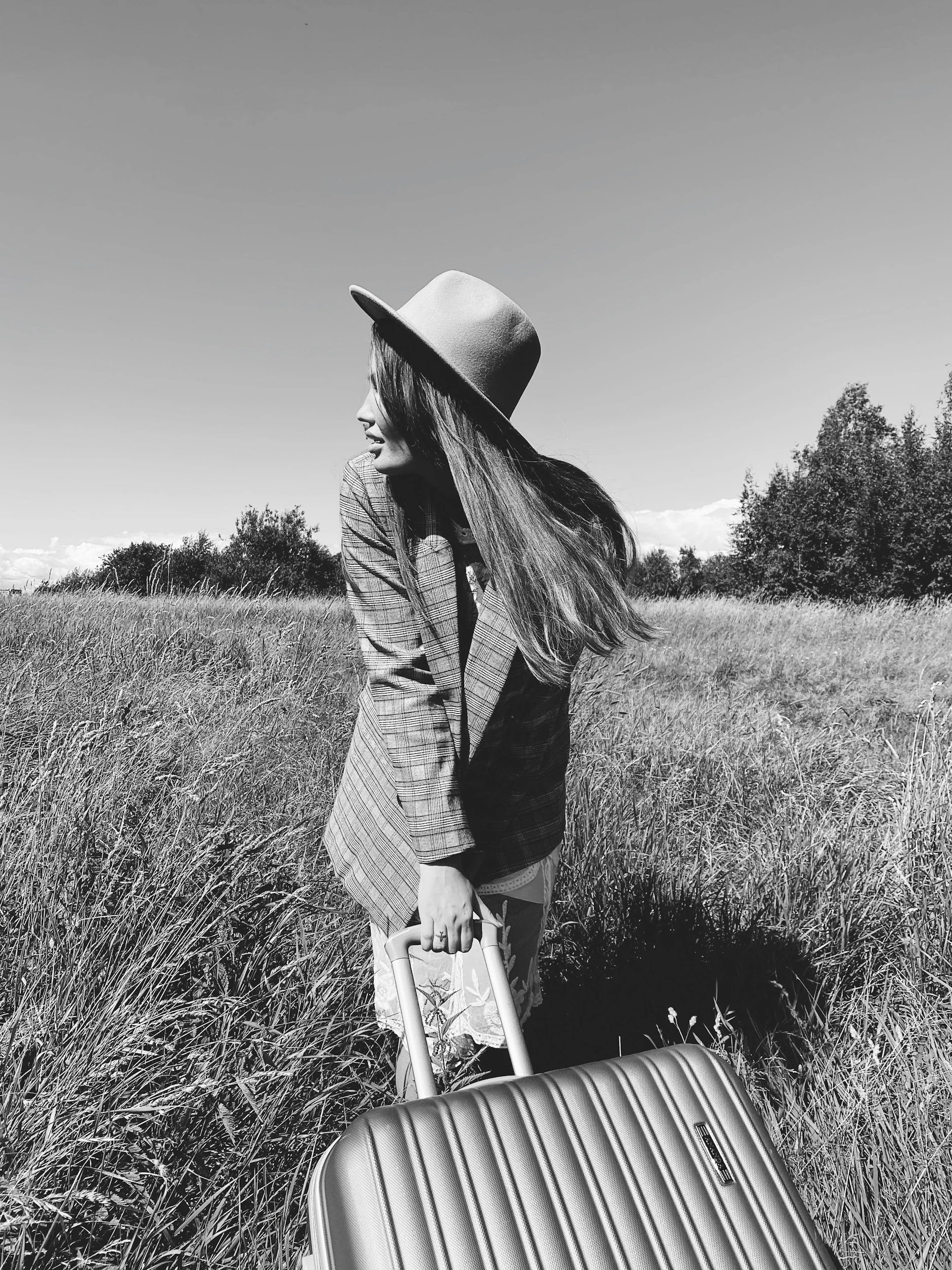 Mujer con sombrero y chaqueta en campo, sosteniendo maleta en mano, vista lateral en paisaje outdoor, en blanco y negro.