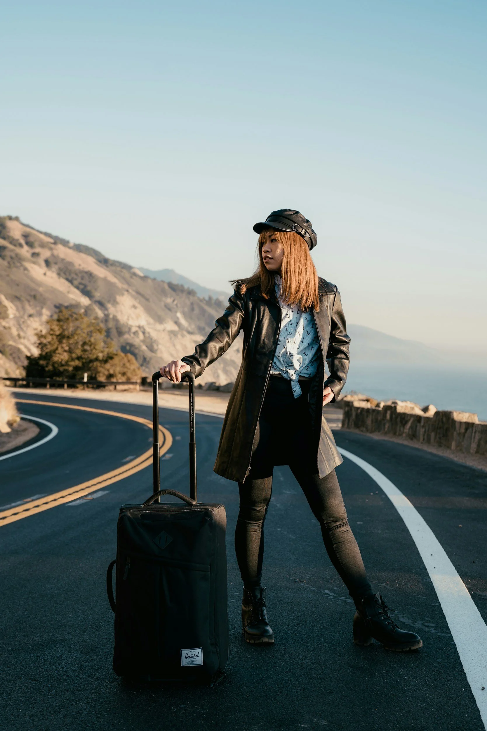 Una mujer con cabello castaño y largo, usando un abrigo negro, pantalones oscuros, botas y gorra negra, de pie en una carretera en un paisaje montañoso con maleta de mano, en un día soleado y despejado.