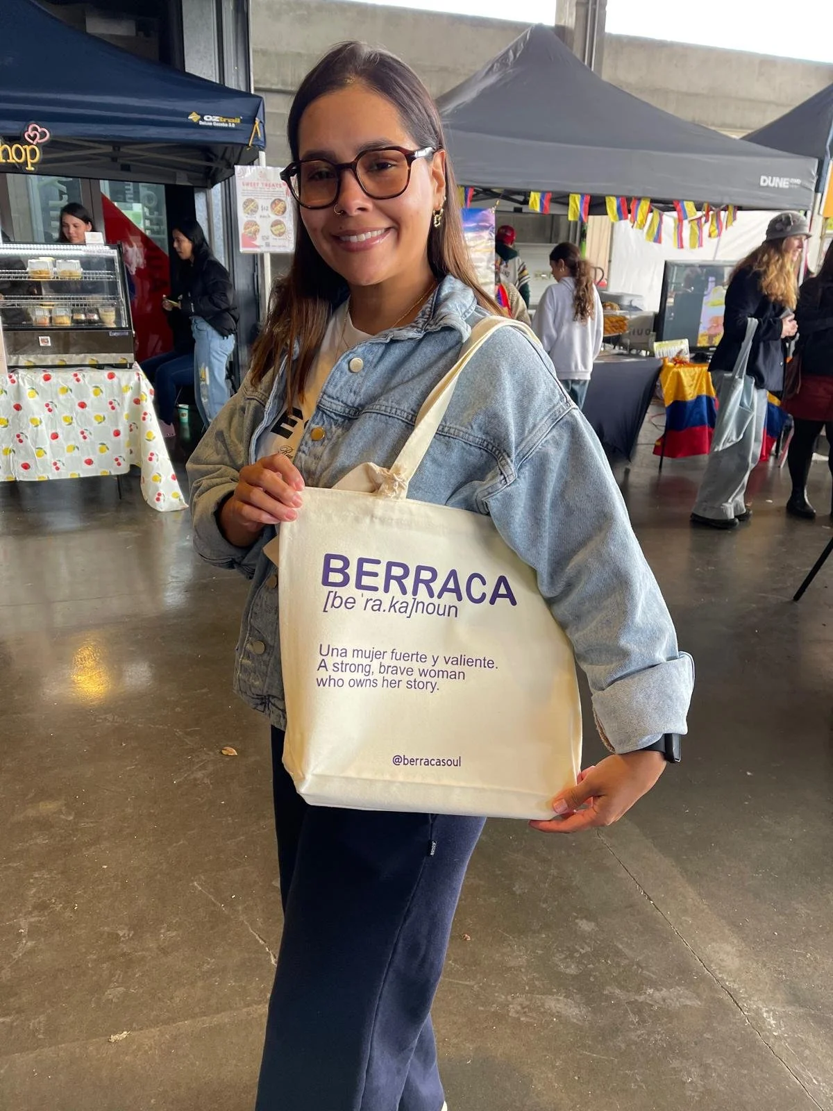Mujer joven con gafas y sudadera de mezclilla sosteniendo una bolsa de tela con la palabra 'BERRACA', que significa una mujer fuerte y valiente, en un evento al aire libre con puestos y otras personas.