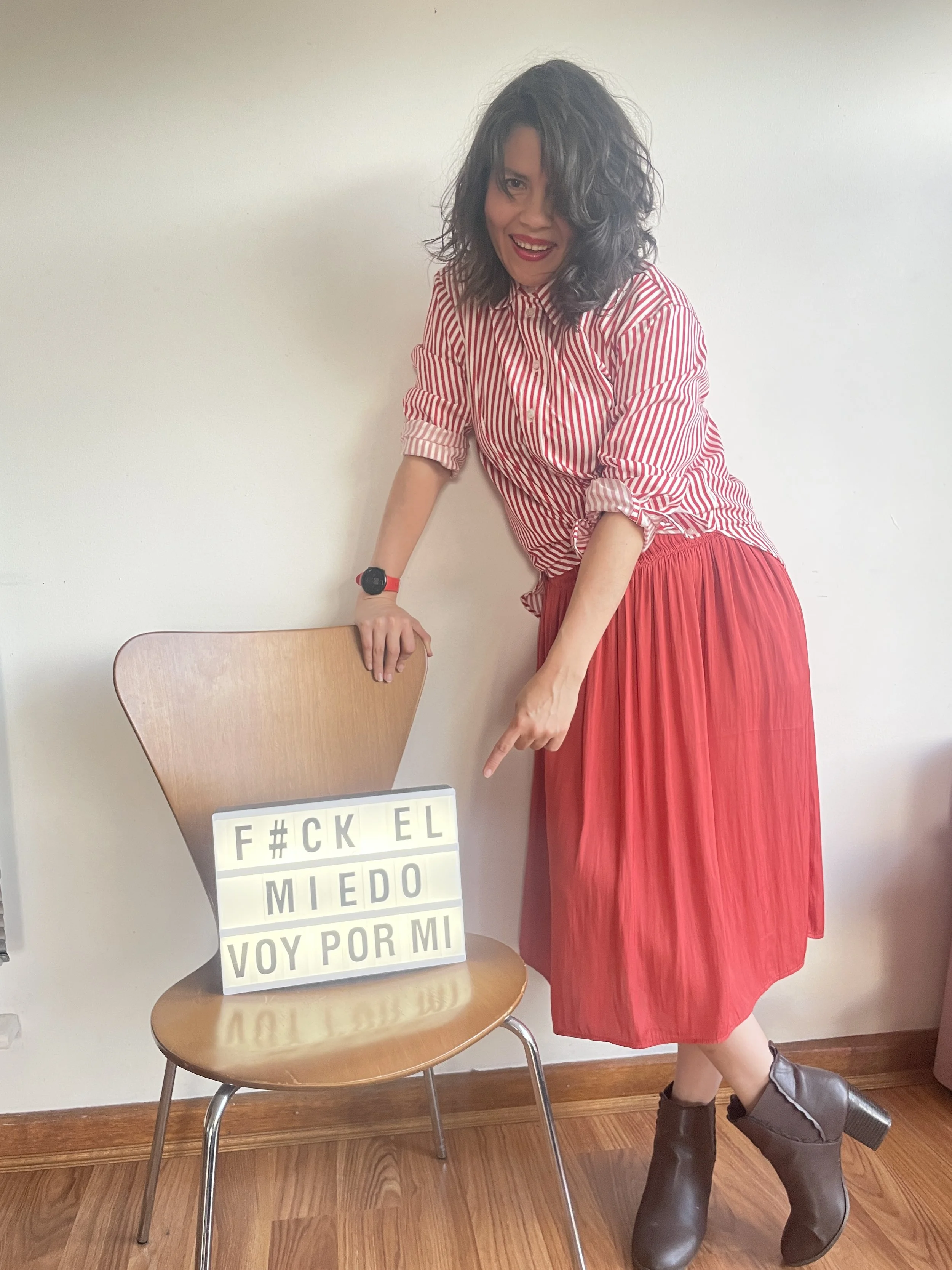 Persona con cabello rizado, camiseta a rayas roja y blanca, falda larga roja, botas negras grandes, señalando una silla con cartel que dice palabras censuradas en español.