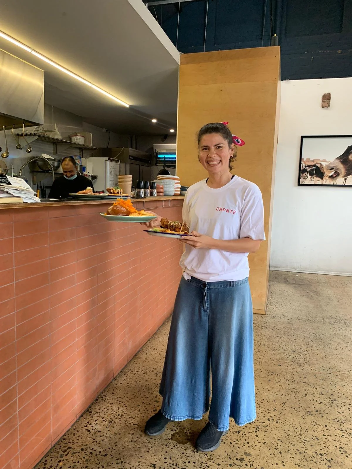 Mujer sonriendo en restaurante, sosteniendo platos con comida, fondo de cocina y pared de ladrillos.