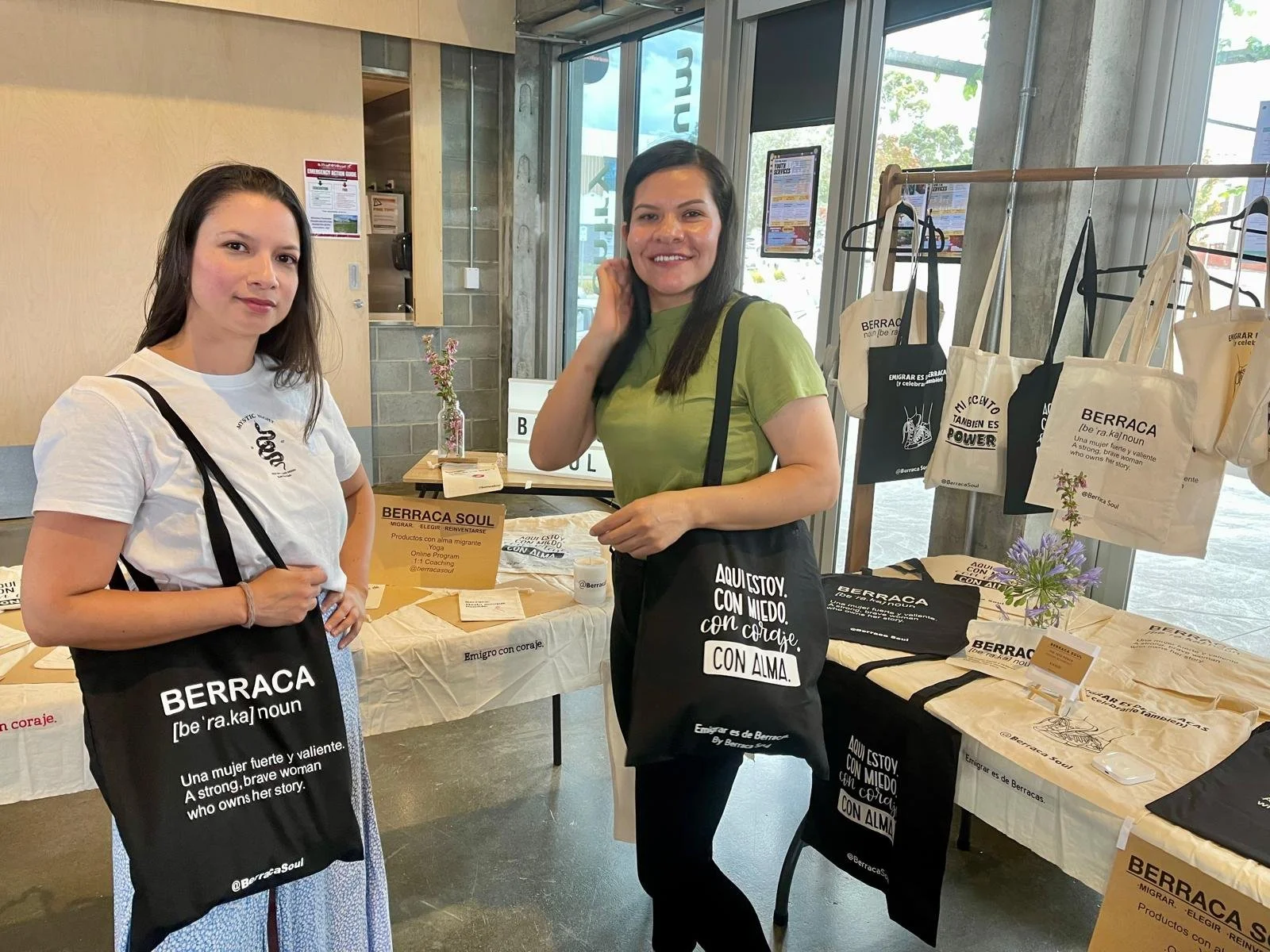 Dos mujeres en un puesto de difusiones, una con blusa blanca y otra con camiseta verde, que llevan bolsos o bolsas con texto sobre la valentía y el empoderamiento de las mujeres, en un ambiente luminoso con mesas y productos en exhibición.