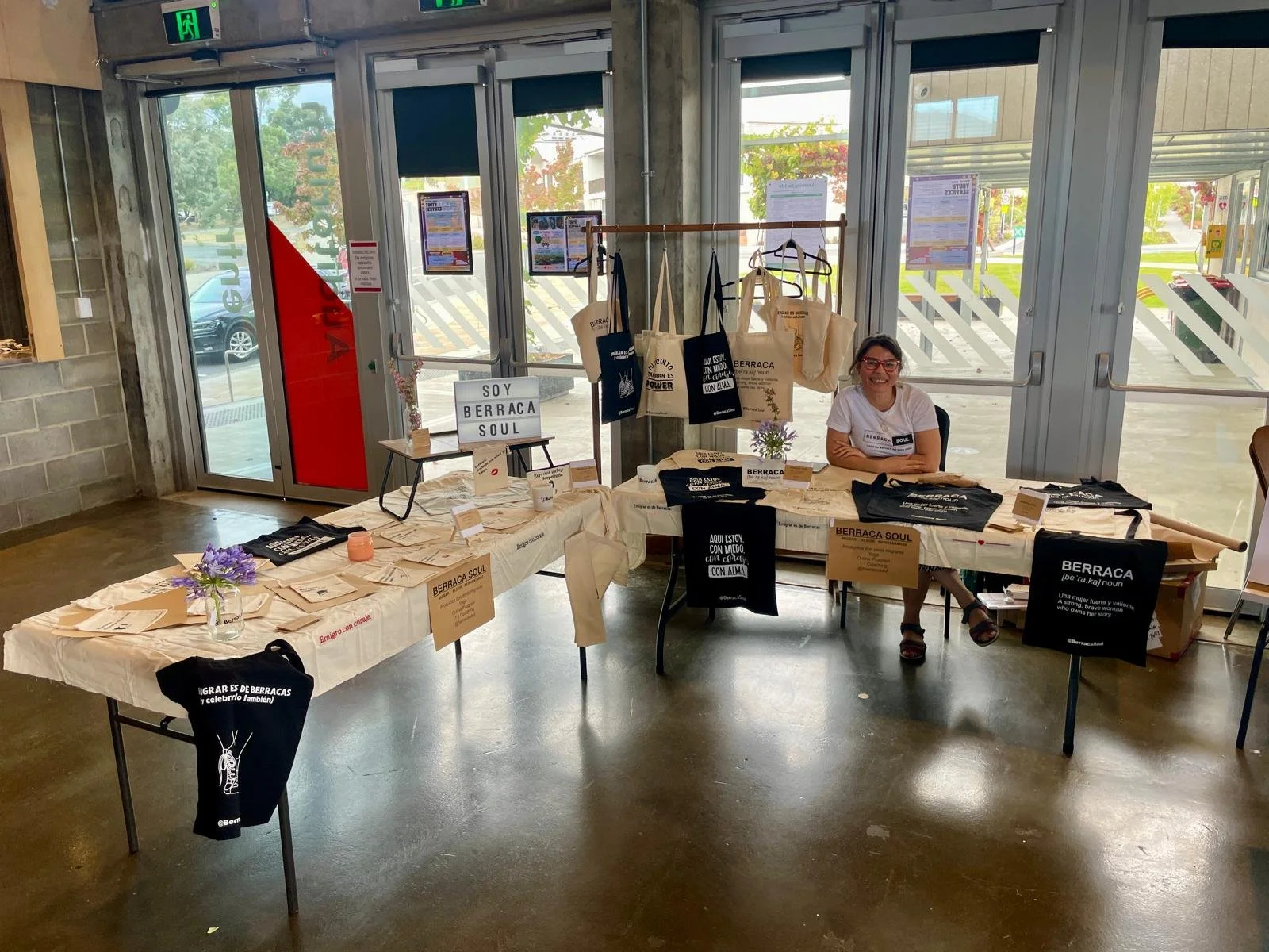 Una mesa con productos y materiales promocionales de Berraca Soul, incluyendo bolsas, carteles y camisetas, en un espacio interior con grandes ventanas y una mujer sonriendo sentada detrás de la mesa.