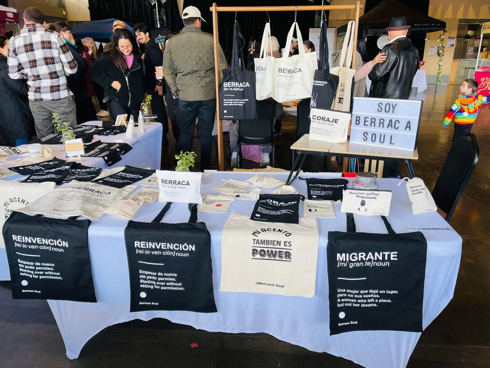 Un puesto con varias bolsas y camisetas con frases en español en un mercado o feria, dedicado a la comunidad migrante. La mesa tiene bolsas de tela blancas y negras, algunas con las frases "Reinvención" y "Migrante". También hay carteles que dicen "Soy BERRACA SOUL" y otros con definiciones y mensajes motivacionales en español.