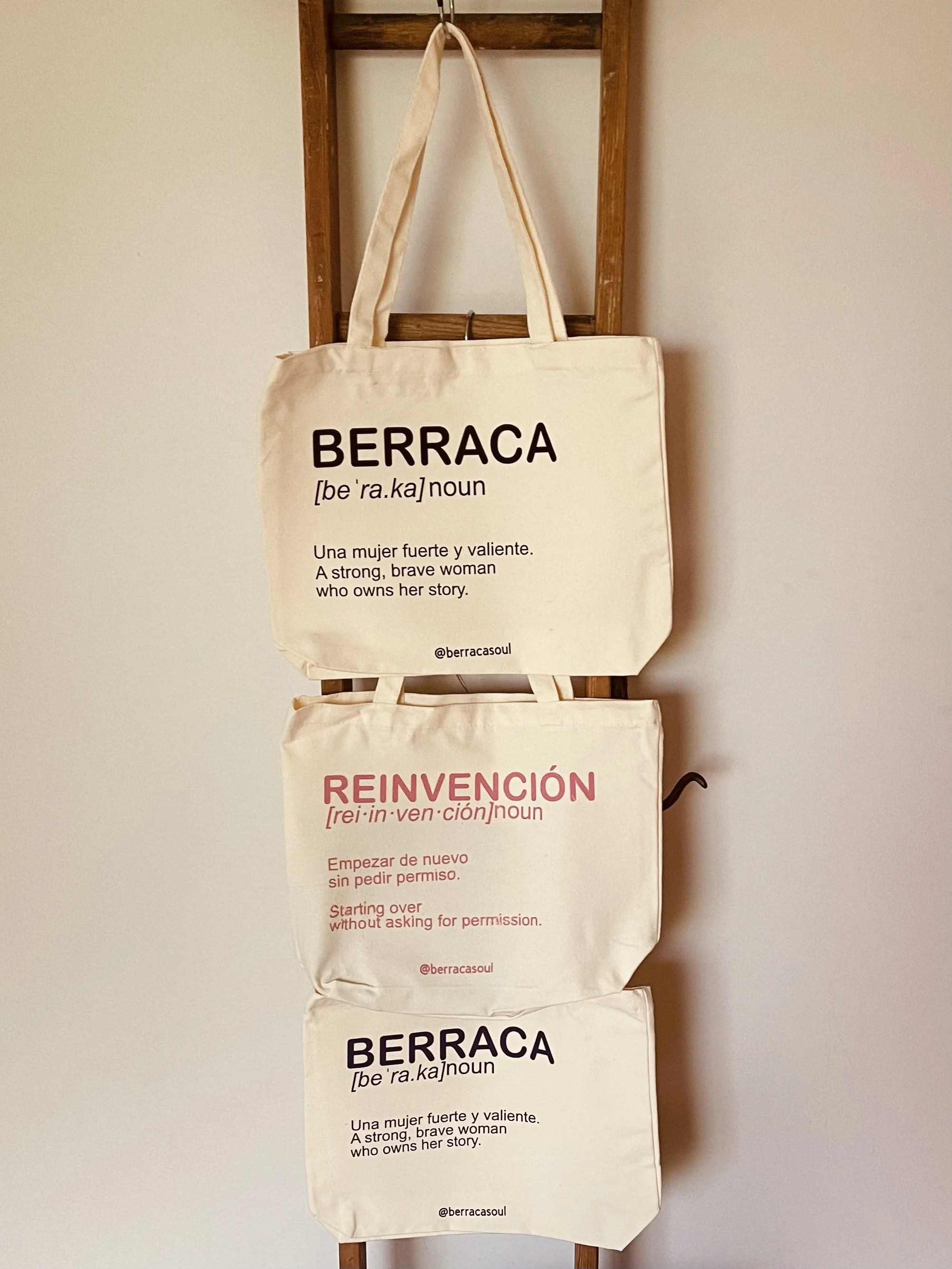 Cuatro bolsas de tela colgadas en una estructura de madera. Cada bolsa tiene palabras en español e inglés relacionadas con el empoderamiento y la valentía de las mujeres, incluyendo definiciones y frases motivacionales.