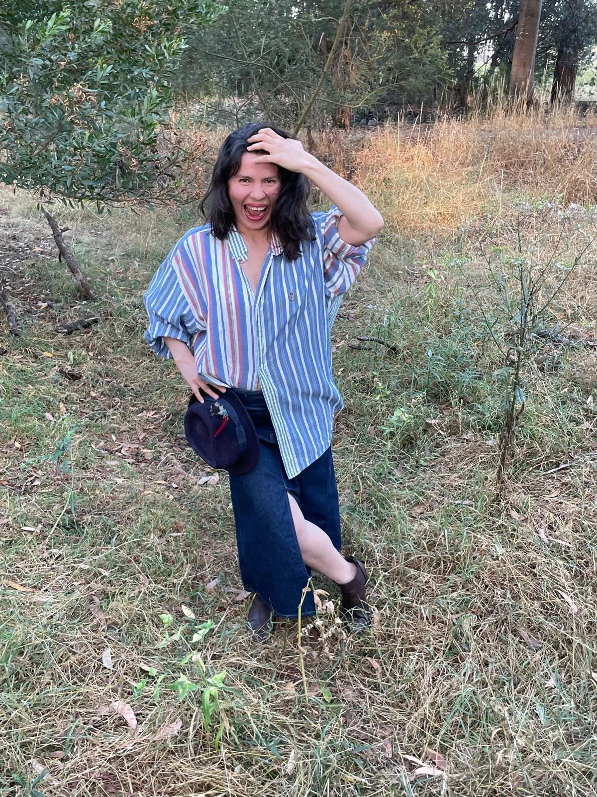 Mujer sonriendo y con expresión de sorpresa, con el cabello negro, vestida con una camisa de rayas azul, blanca, y roja, pantalones oscuros, en un área de hierba y árboles.