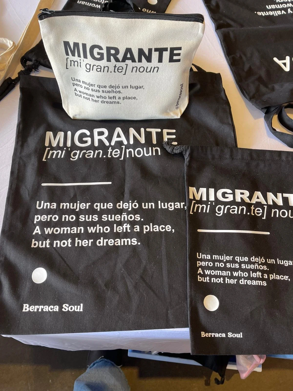 Lonas negras y una bolsa de tela blanca con la palabra 'MIGRANTE' y su definición en inglés y español. La definición en español dice: 'Una mujer que dejó un lugar, pero no sus sueños. A woman who left a place, but not her dreams.'