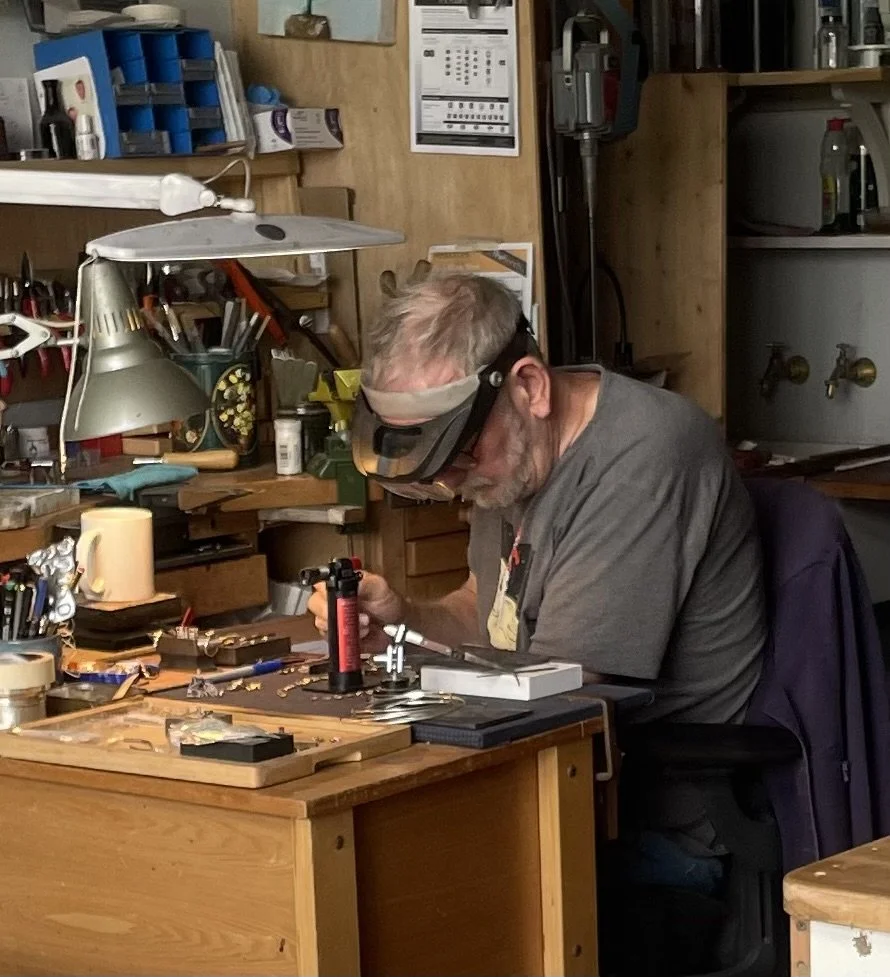 A man wearing a magnifying visor and safety glasses working on jewelry at a workbench in a workshop.