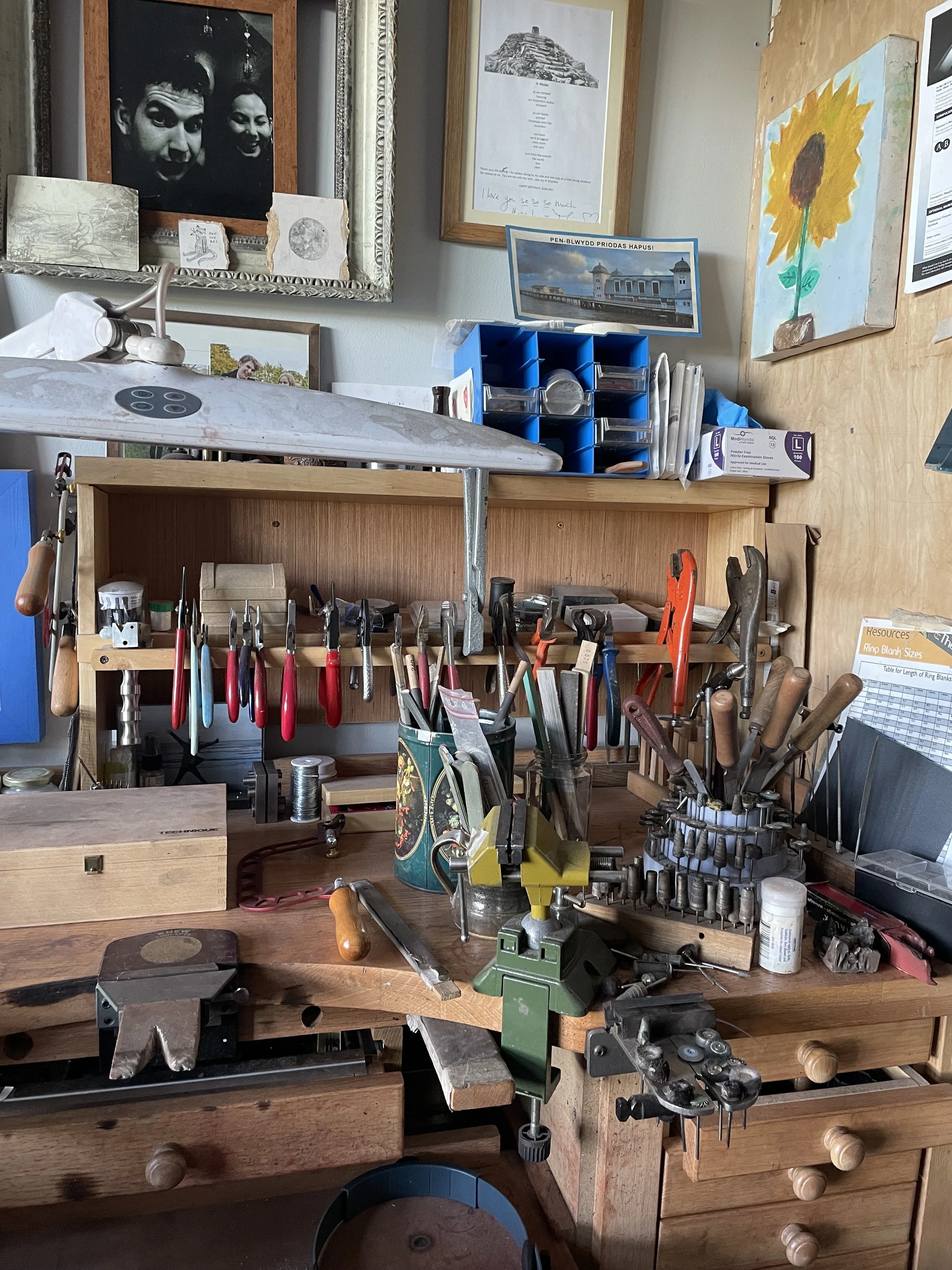 A woodworking workbench with various hand tools including pliers, screwdrivers, chisels, and clamps, surrounded by framed pictures and notes on the wall.