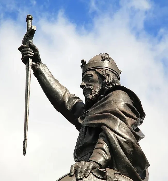A bronze statue of a medieval King Alfred holding a sword high in his right hand, with a clouded sky in the background.
