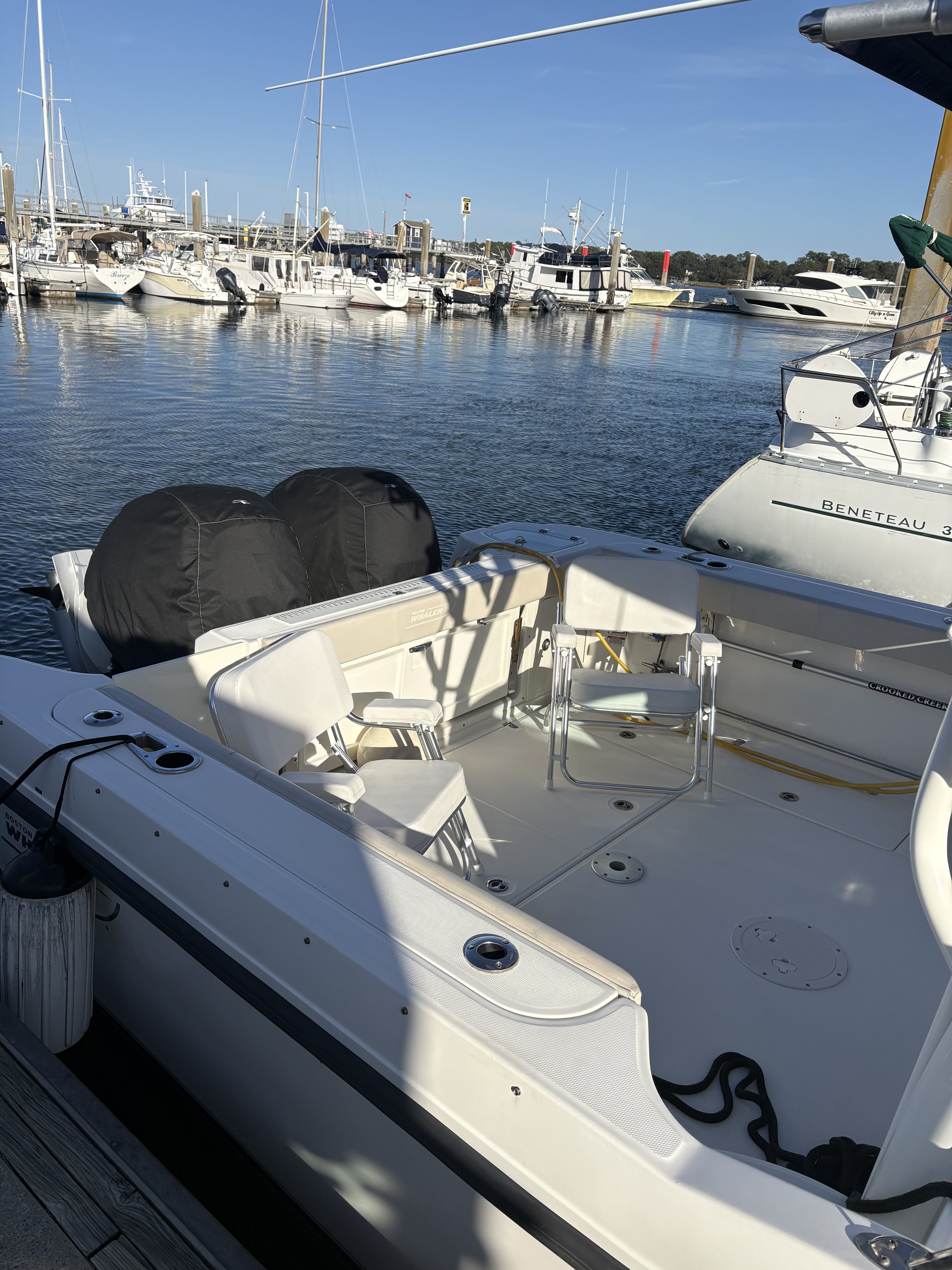 View of a marina with multiple boats docked, including a close-up of a white boat's deck with two outboard motors, chairs, and seating area, clear blue sky.