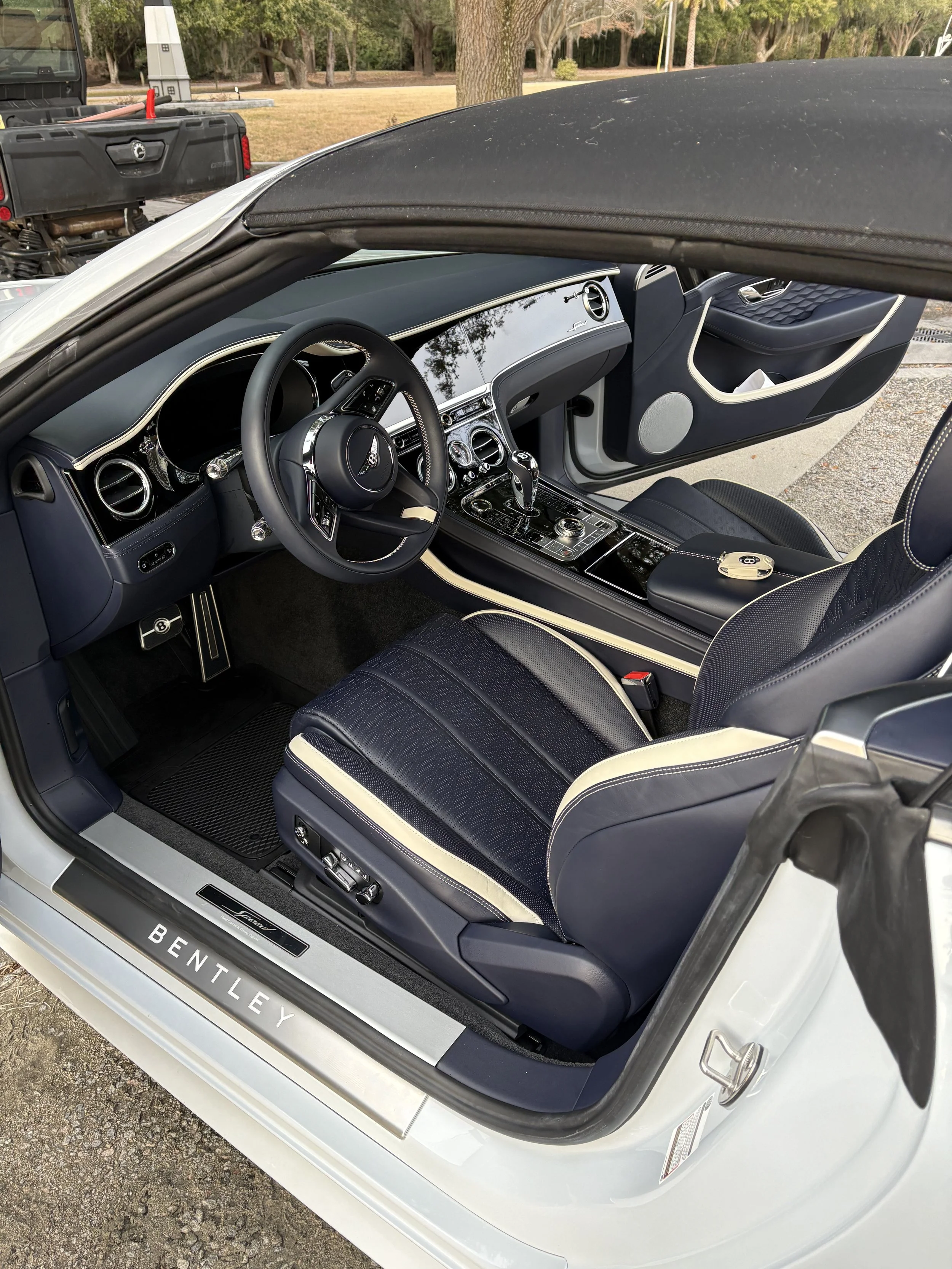 Interior of a white Bentley convertible with black and cream leather seats, dashboard, and center console, outside on a paved area with trees in the background after a detail in beaufort