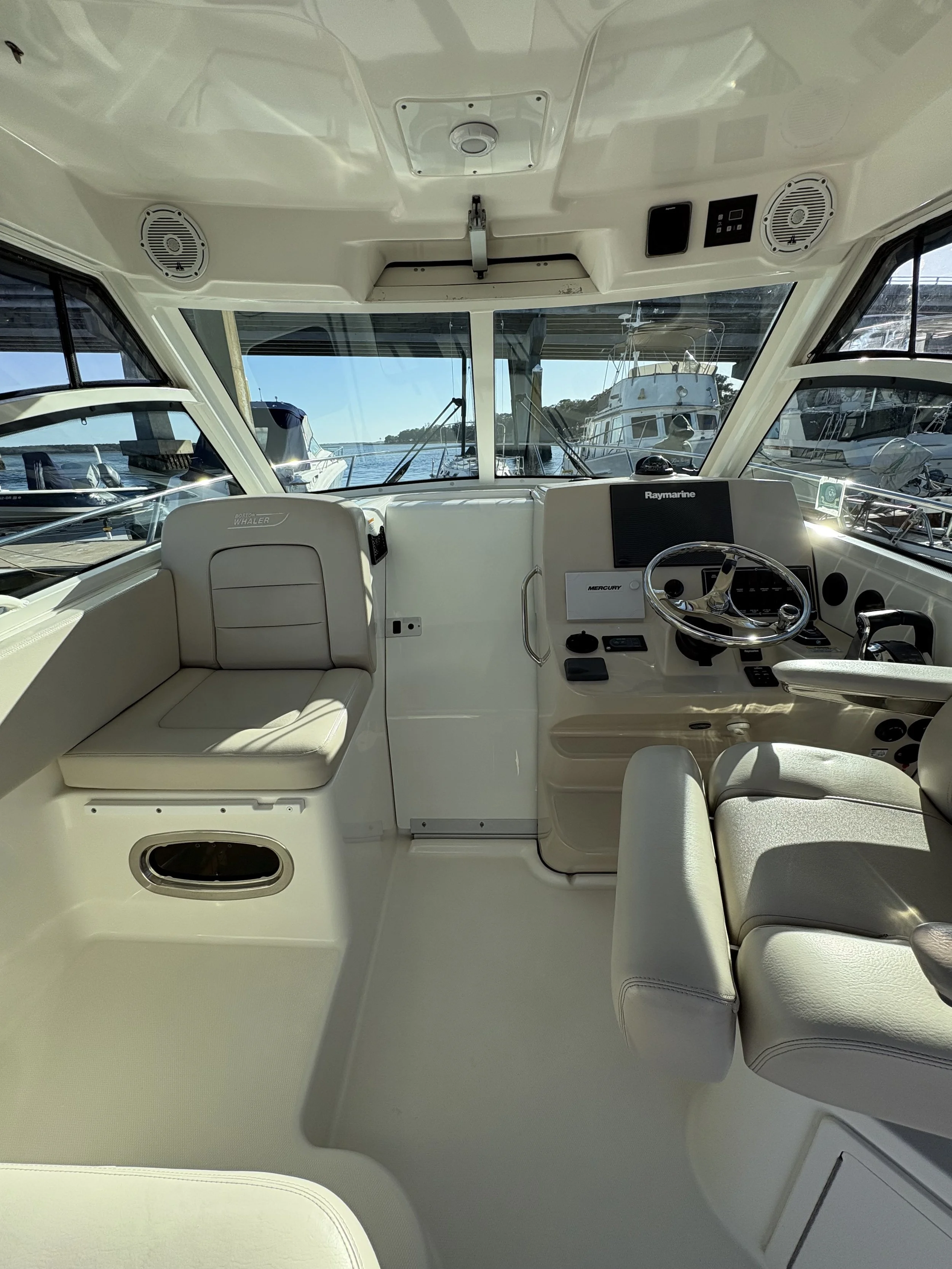 The interior of a boat cockpit with cream-colored seating, steering wheel, and navigation equipment, with view through windows of docked boats and water outside.