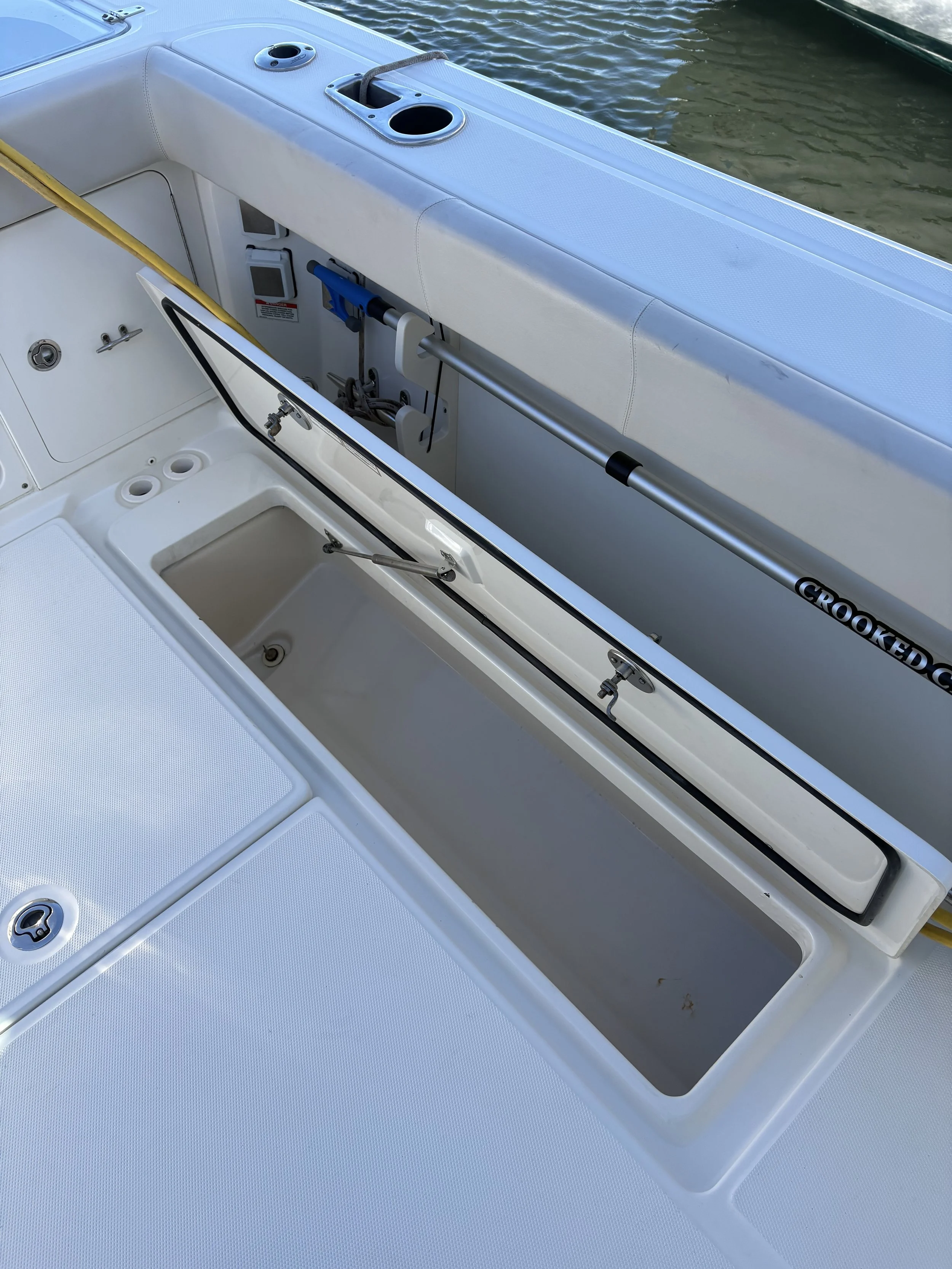 Open storage compartments on a boat with various tools and equipment, including a fishing rod and a croaked cushioned railing, on calm water.