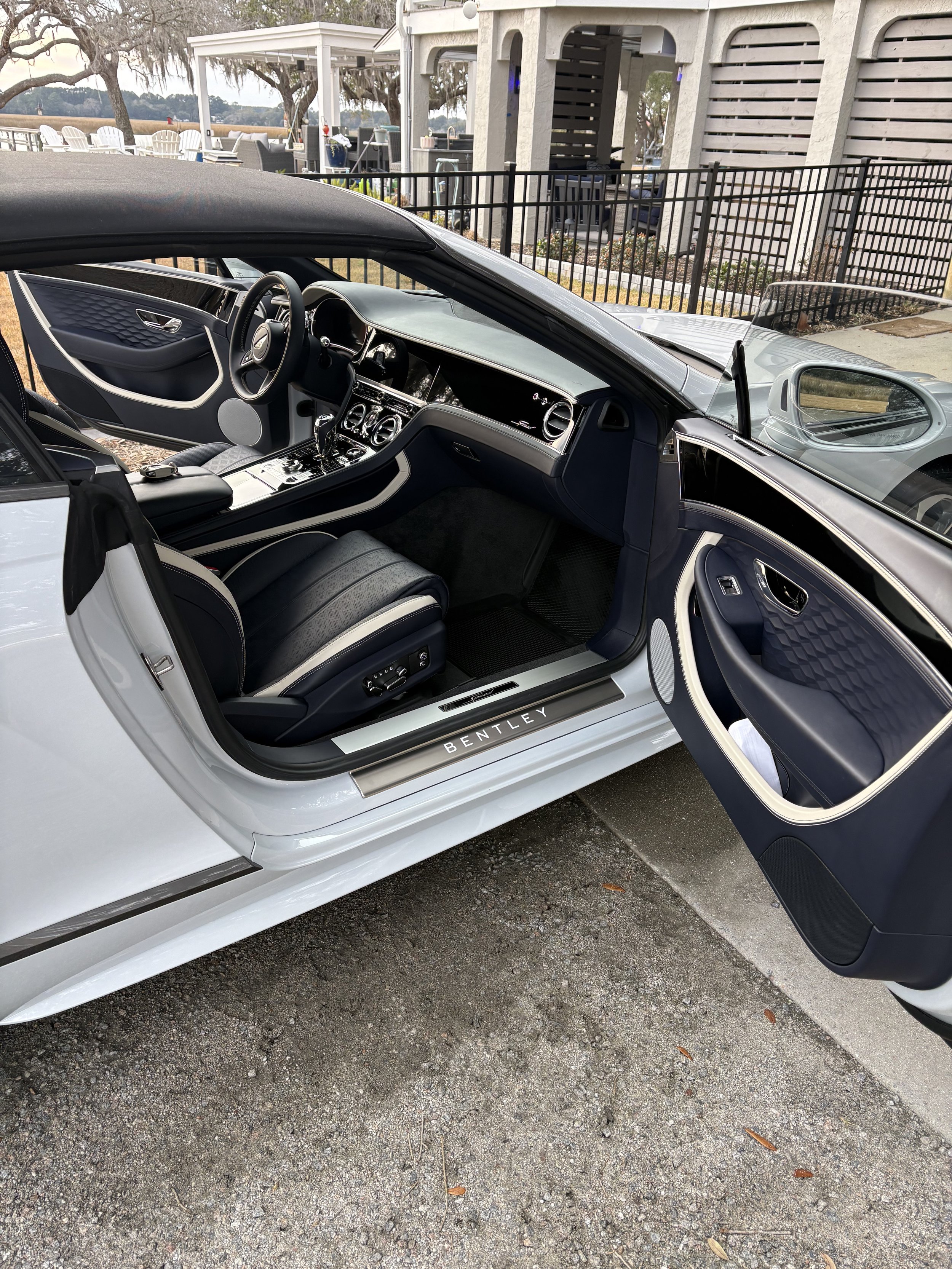 Interior of a luxury Bentley sports car with a black and white color scheme, showing the dashboard, steering wheel, and seats, parked outdoors near a waterfront with chairs and fencing visible in the background after a detail in beaufort
