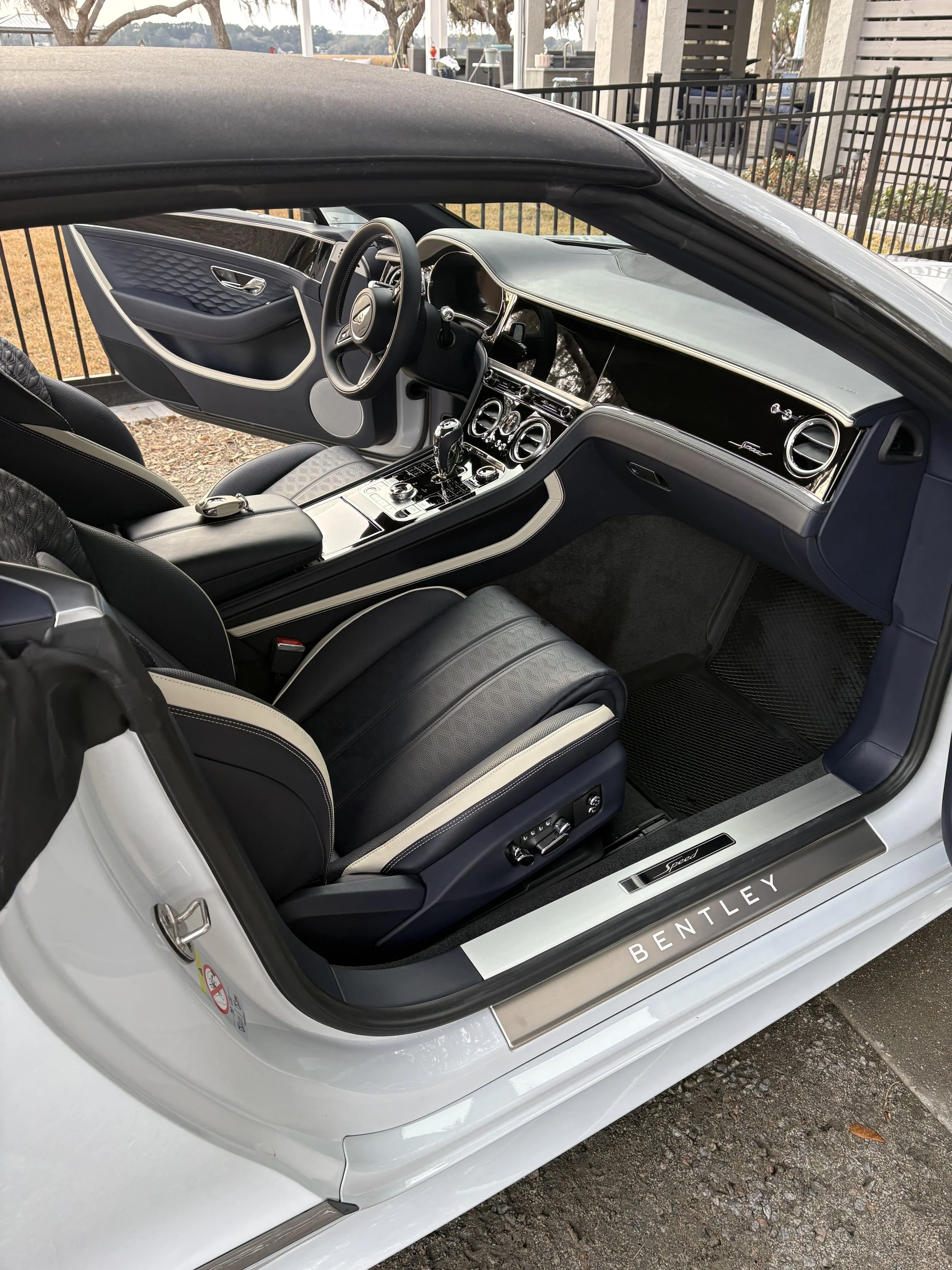 Interior of a luxury white Bentley coupe with black and beige leather seats, a sleek dashboard, and a gear shifter, parked outdoors near a black fence.
