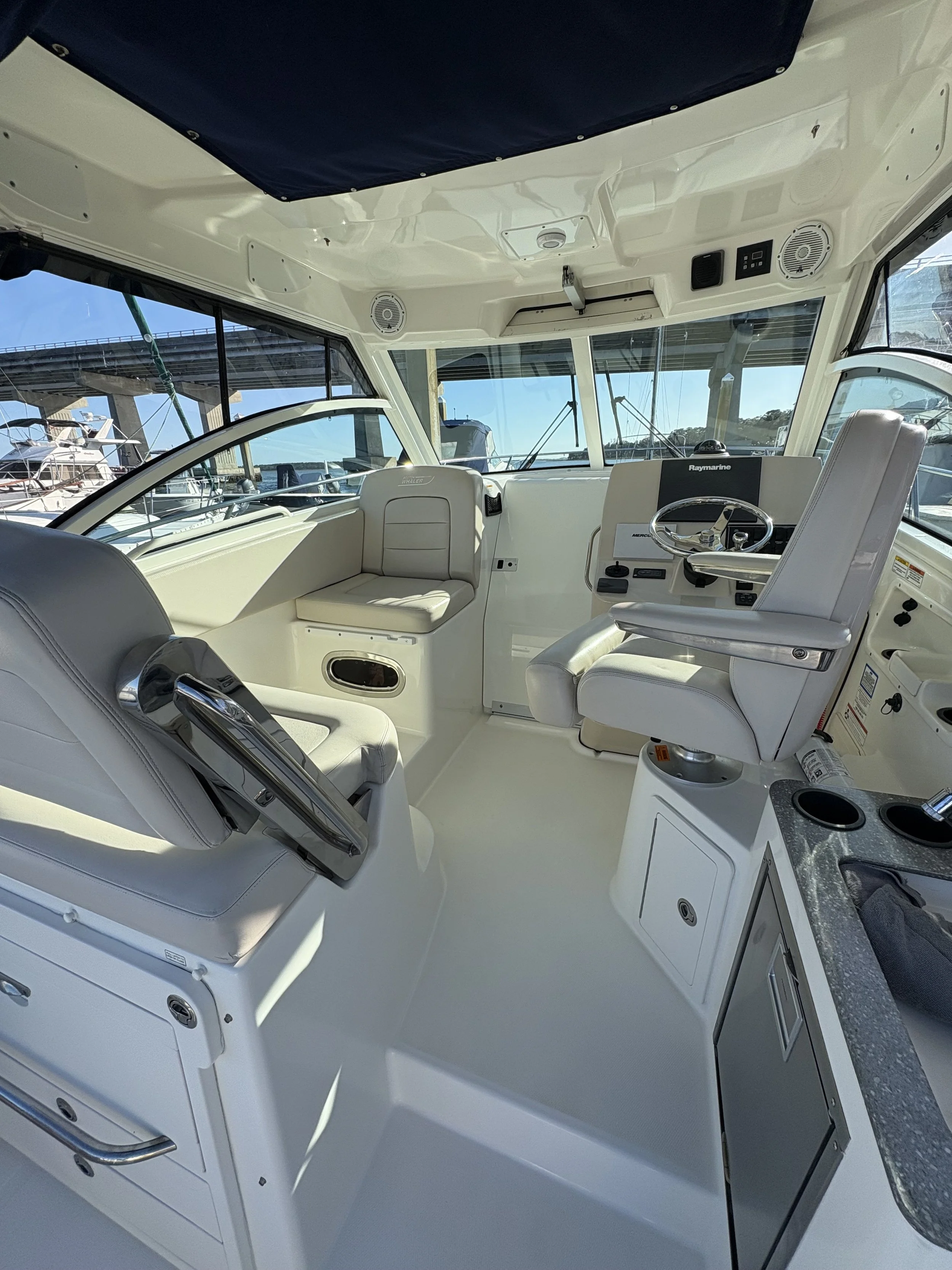 Interior of a white yacht's helm station with leather seats, steering wheel, navigation equipment, and large windows showing nearby boats and water.