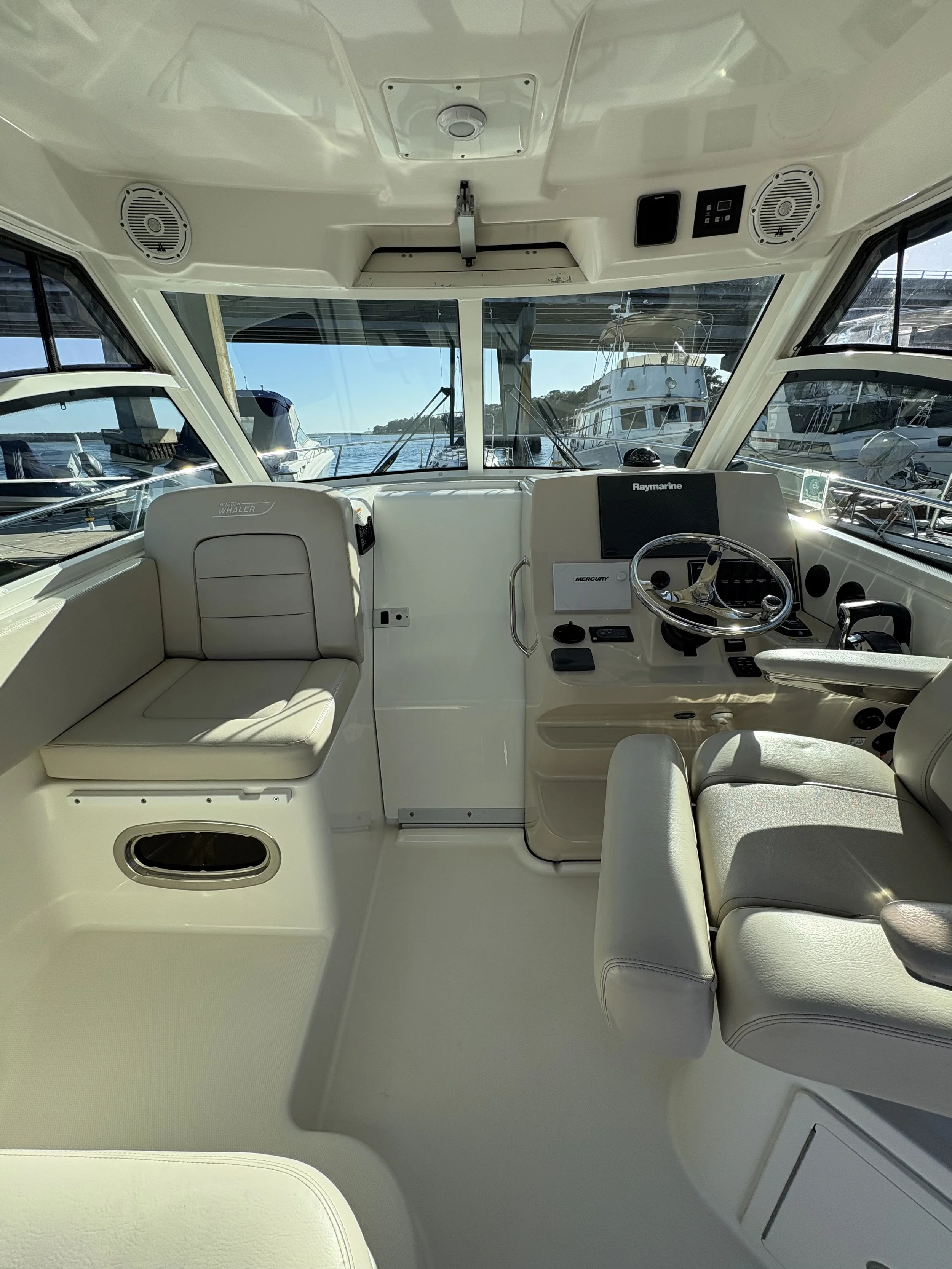 Interior of a boat's helm station with navigation equipment, steering wheel, and seating, overlooking a marina with other boats.