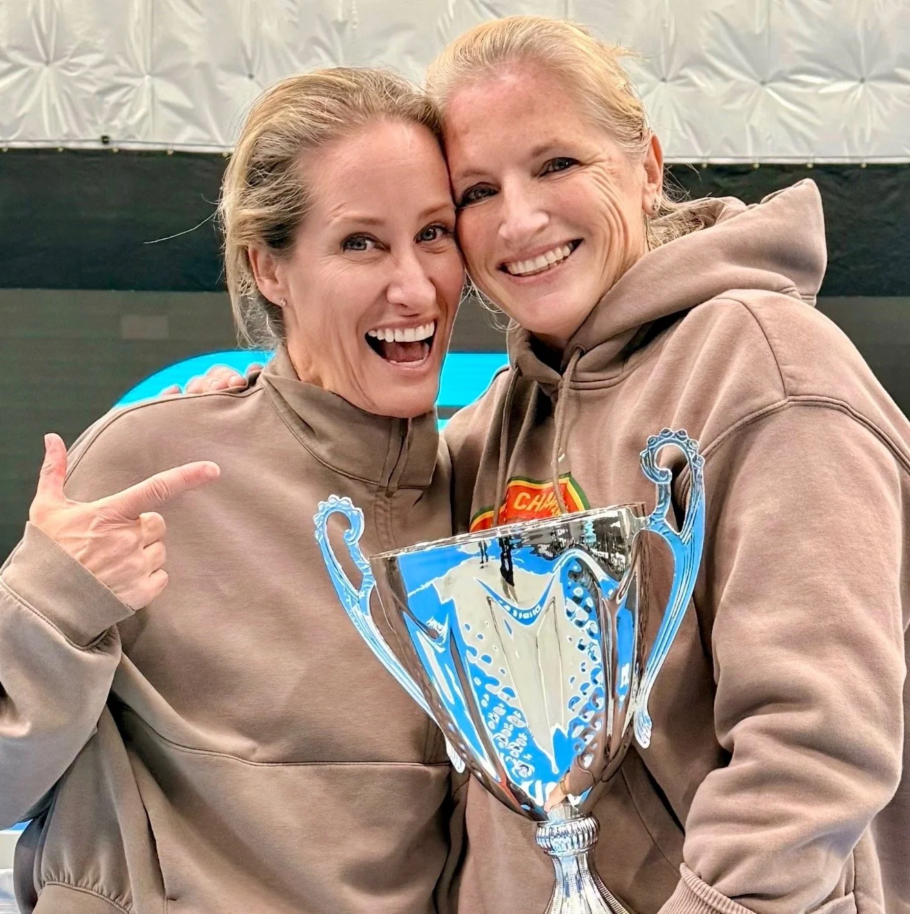 Two smiling women wearing matching beige sweatshirts, one pointing at a large silver trophy they are holding, celebrating a victory or achievement.
