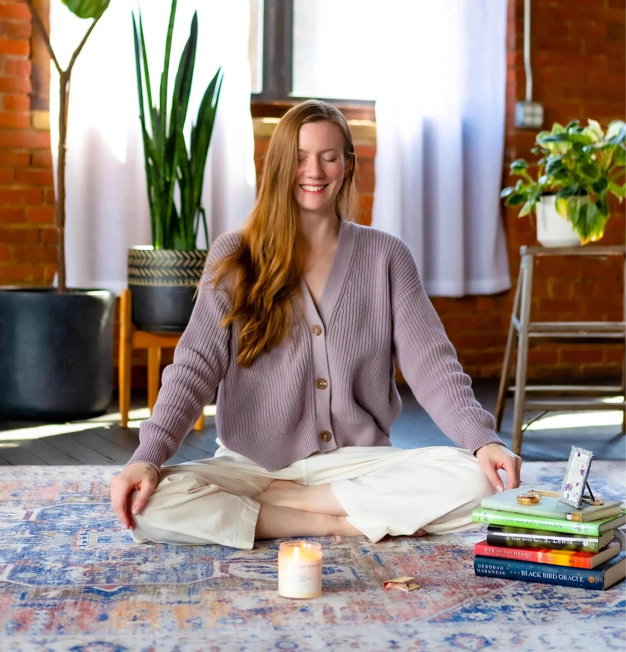 A woman with long red hair sits cross-legged, smiling in meditation, as a white candle burns