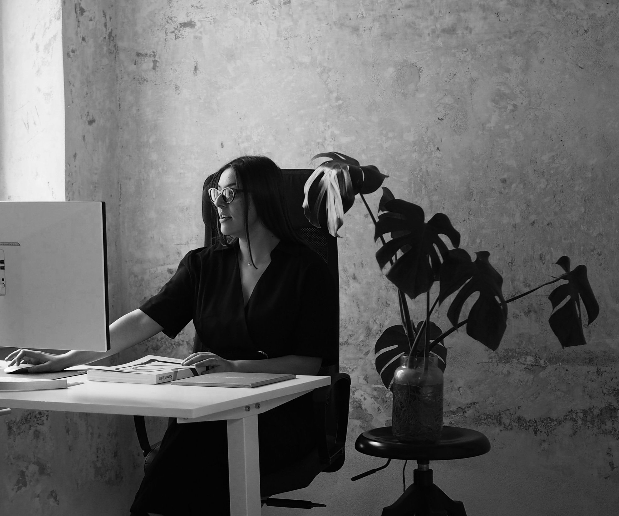A woman with long dark hair and glasses sitting at a desk, working on a computer in a minimalistic office space. There is a potted plant on a stool next to her.